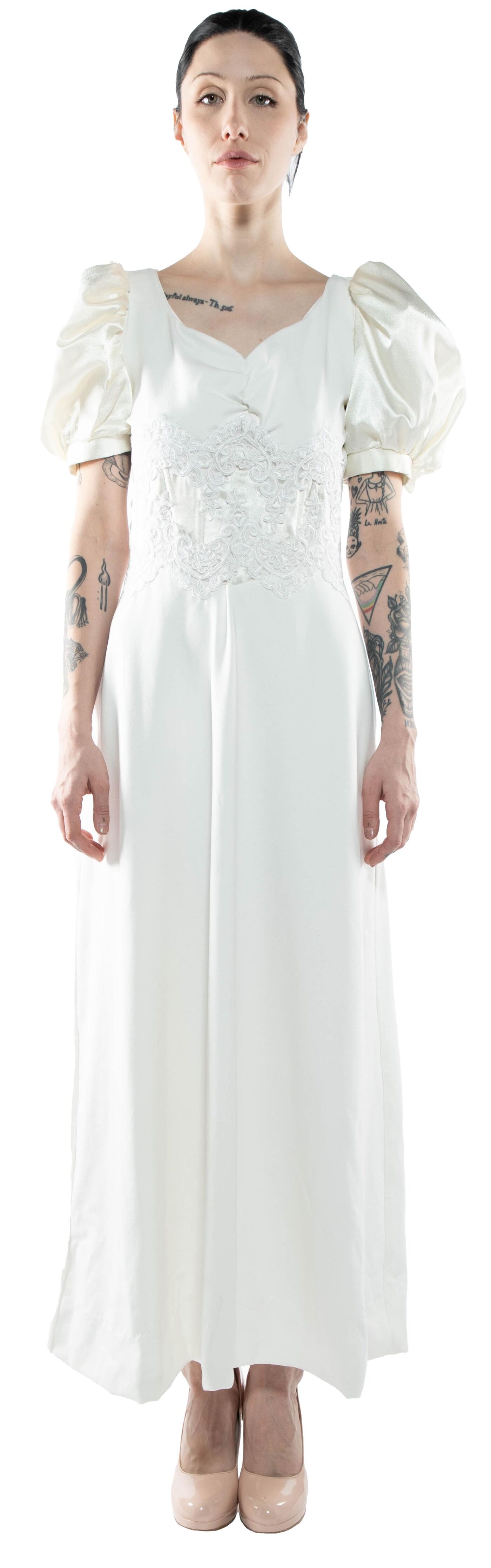 A full shot of a woman with tattoos on her arms wearing a white dress with puffy sleeves and lace bodice, standing against a white background.