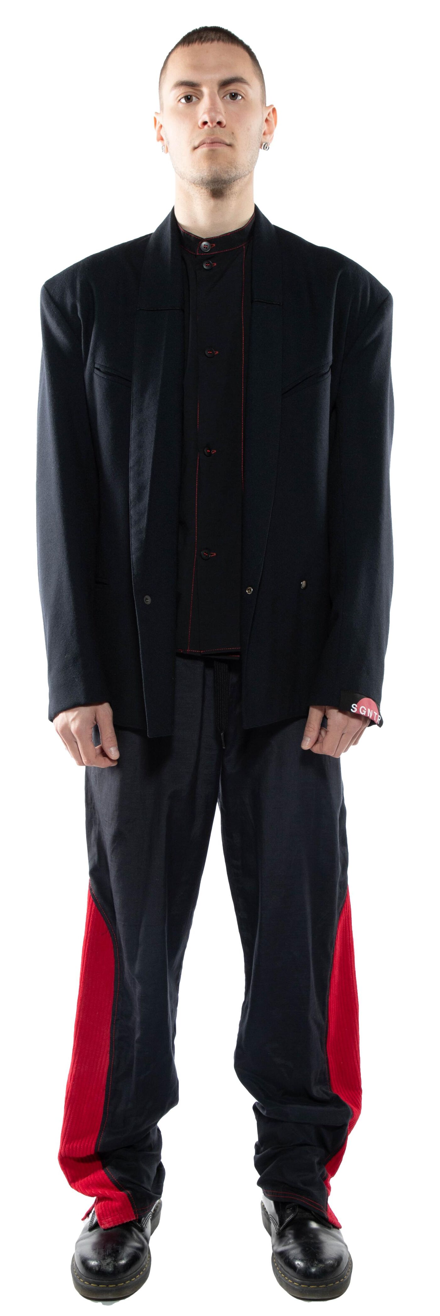 A man with short brown hair is standing against a white background wearing a black blazer, a dark brown collared shirt, and black pants with red stripes down the sides, and black shoes.