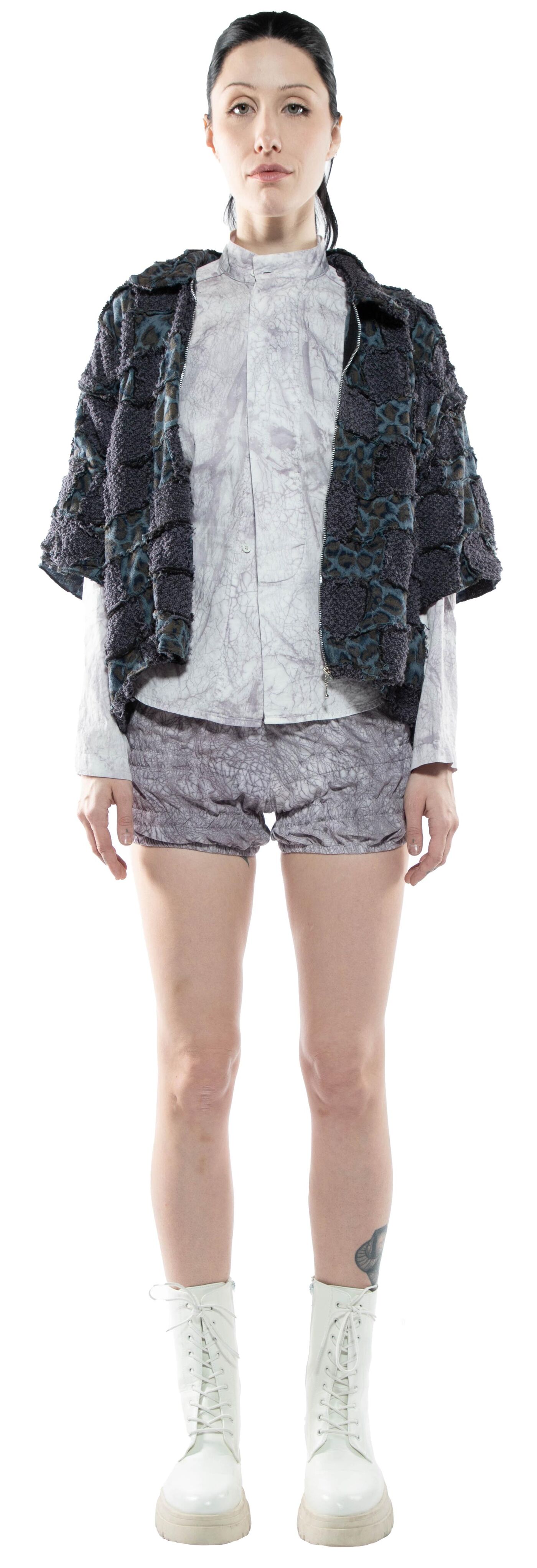 A woman with dark hair and a neutral expression stands against a white background, wearing a dark gray textured jacket, a light gray shirt, light gray shorts, and white lace-up boots.