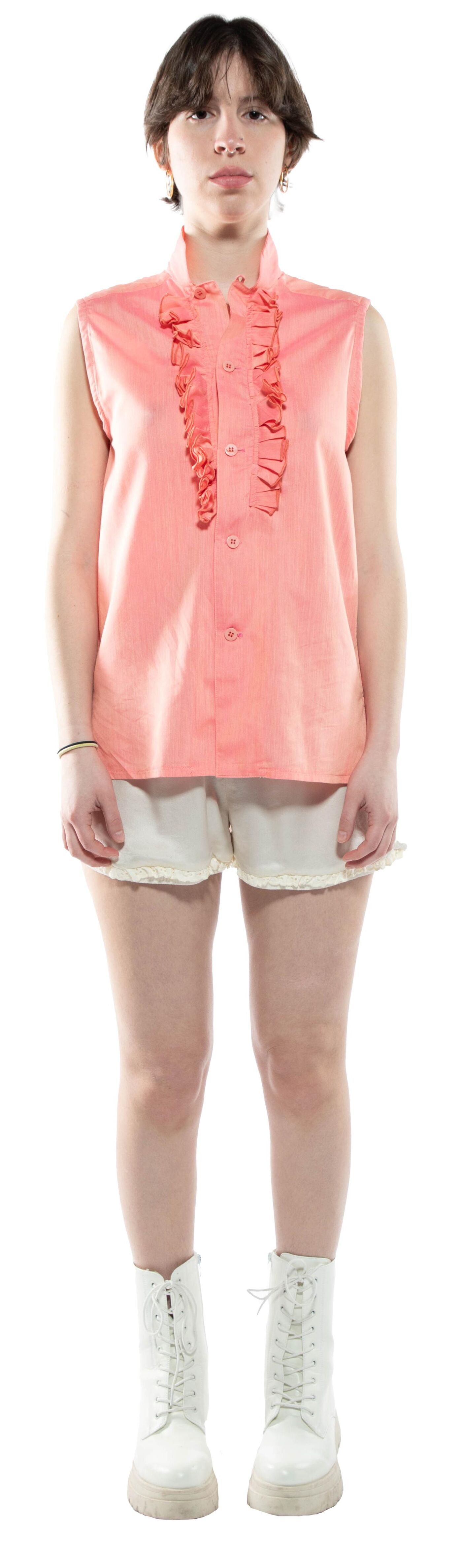 A young woman with short brown hair stands against a white background wearing a coral sleeveless blouse with ruffles, off-white shorts, and white combat boots.