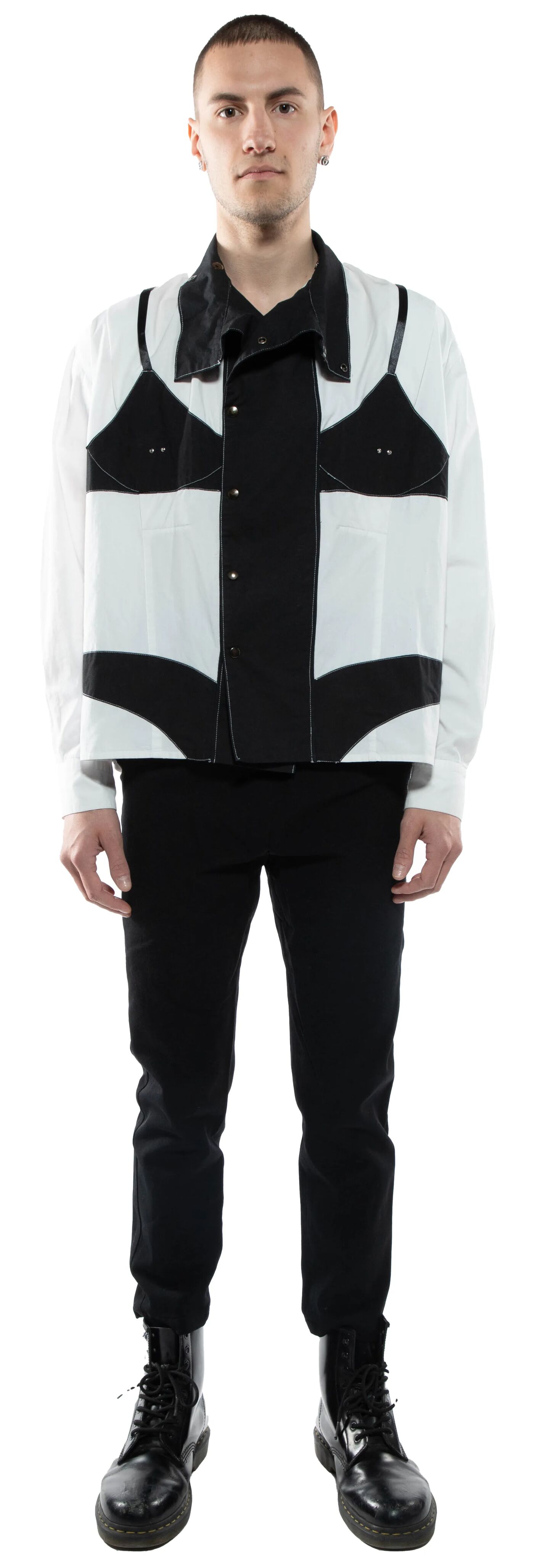A young, light-skinned man with short brown hair stands against a white background wearing a black and white button-up shirt, black pants, and black combat boots.