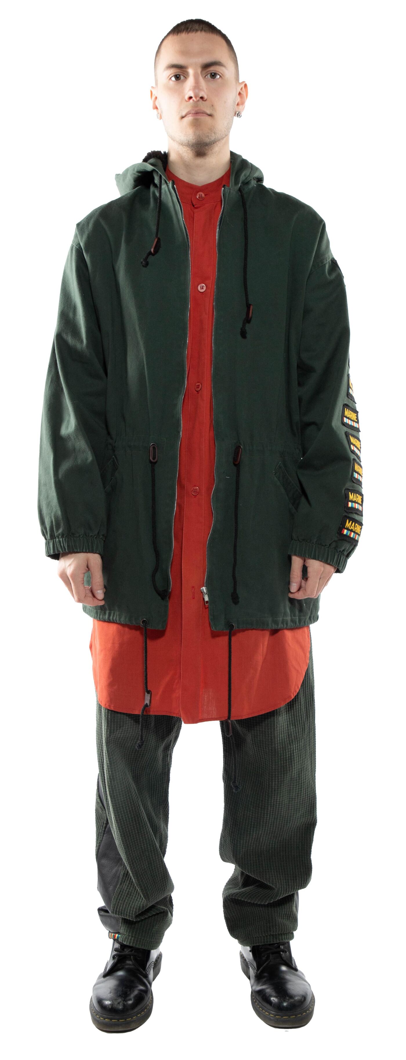 A man stands against a white background wearing a dark green hooded jacket over an orange shirt and dark green pants, with black shoes.