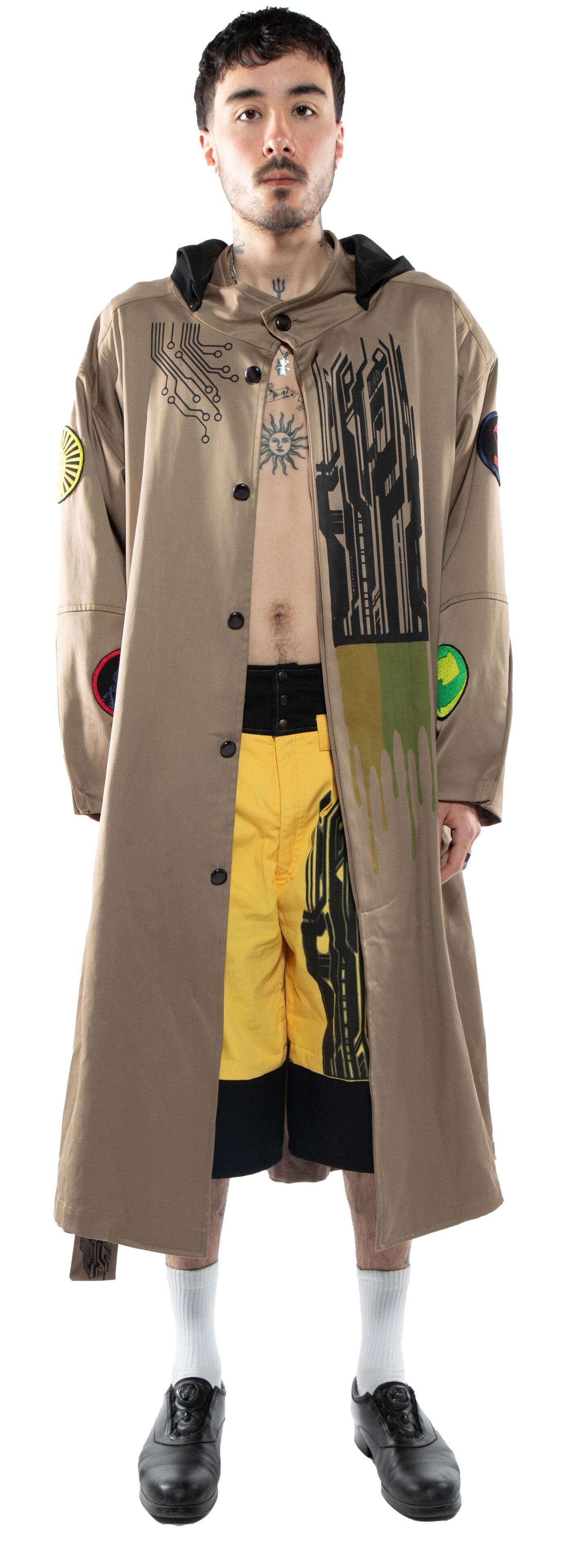 A full shot shows a man in a long taupe trench coat with graphic designs, yellow shorts, black socks, and black shoes standing against a white background.