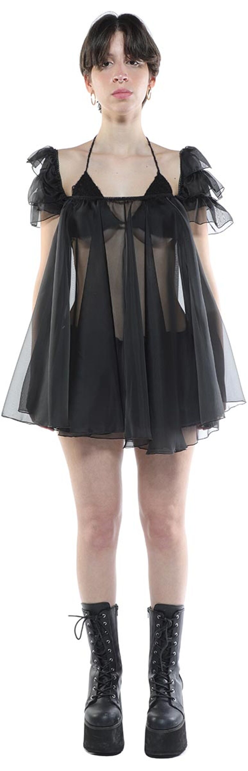 A young woman with short dark hair wears a sheer black mini dress and black platform boots.