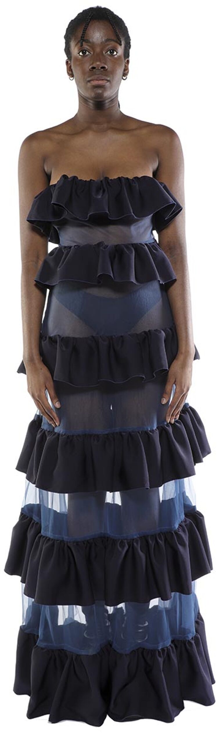 A dark-skinned woman with short black hair stands against a white background wearing a layered, ruffled, strapless maxi dress that is primarily black with translucent blue panels.