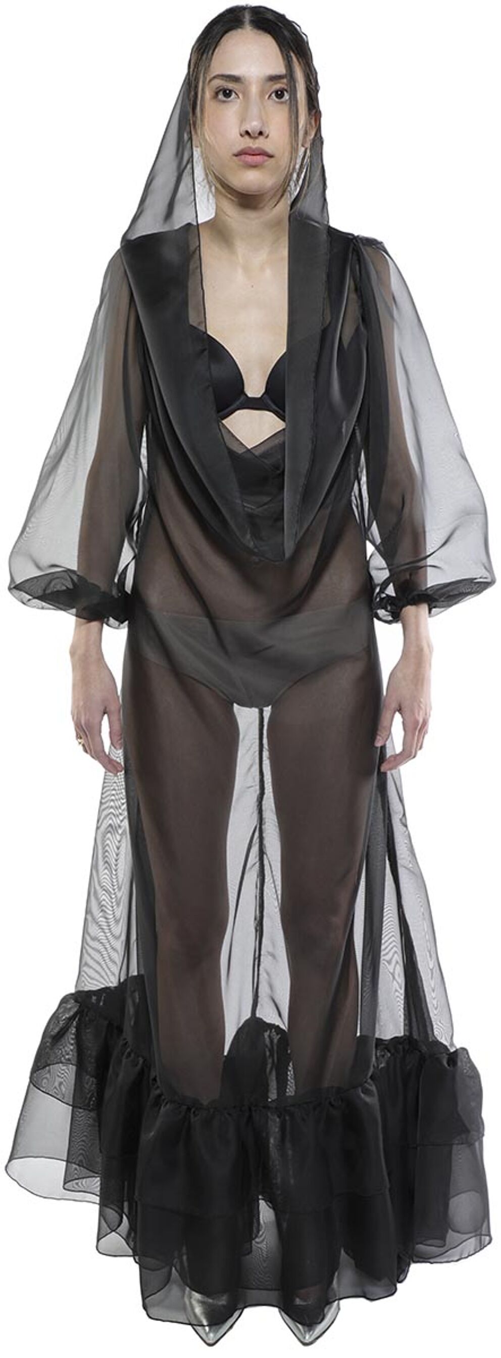 A model wears a sheer black long-sleeved gown with a ruffled hem and a hood, posing against a white background.