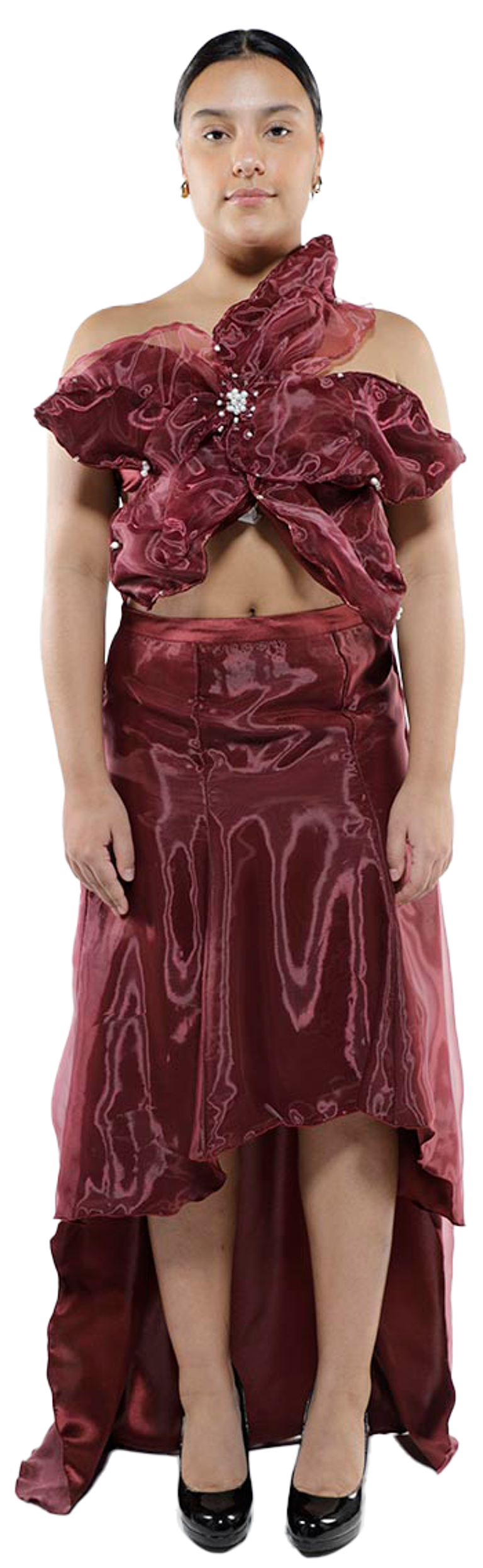 A young woman with tan skin and dark hair, wearing a maroon high-low dress with a ruffled, one-shoulder bodice and embellished with rhinestones, poses against a white background while wearing black heels.
