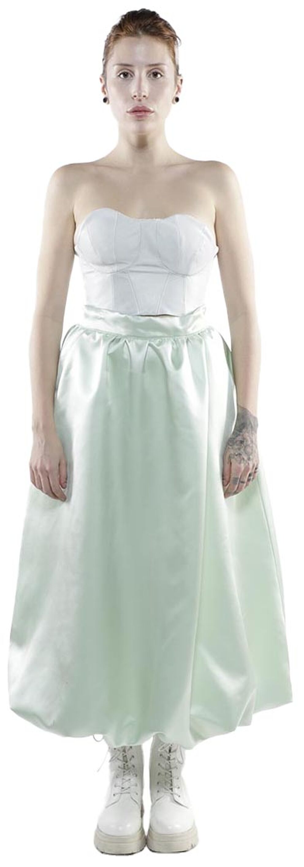 A fair-skinned woman with short reddish-brown hair stands against a white background wearing a light green satin skirt and a white strapless top, paired with white combat boots.