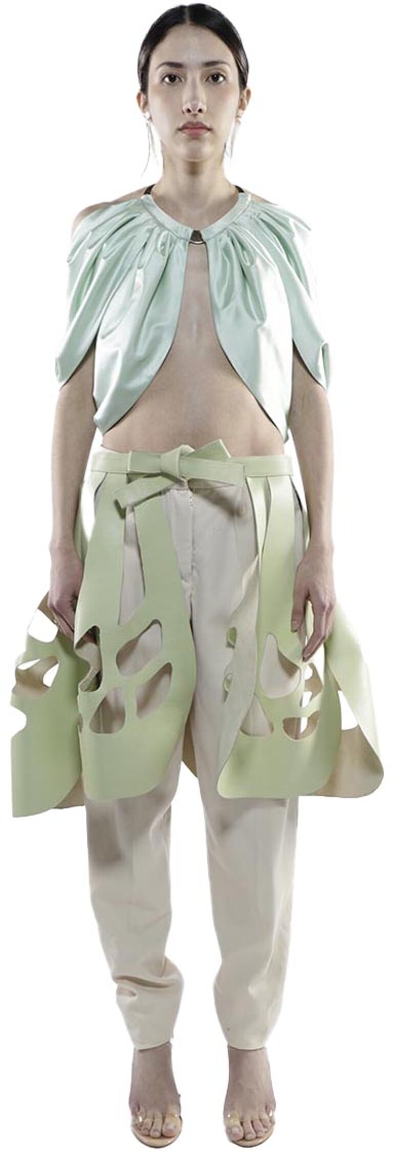 A woman with dark hair pulled back wears a pale green cropped bolero and matching pants with cut-out details, paired with clear heeled sandals.