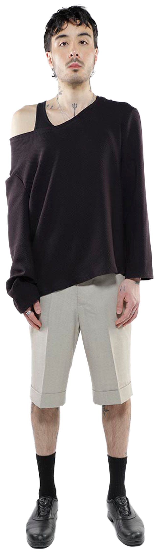 A young man with dark hair and tattoos stands against a white background wearing a black, off-the-shoulder sweater, beige shorts, black socks, and black shoes.
