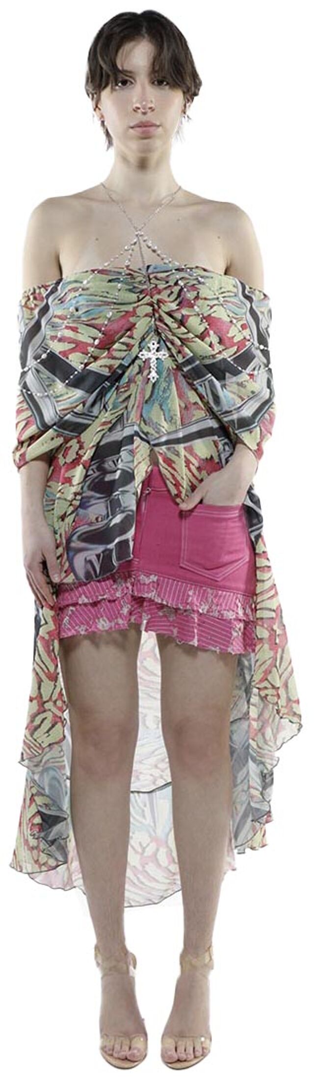 A woman with short brown hair wears a multicolored, off-the-shoulder shawl over a pink mini skirt and clear high heels.