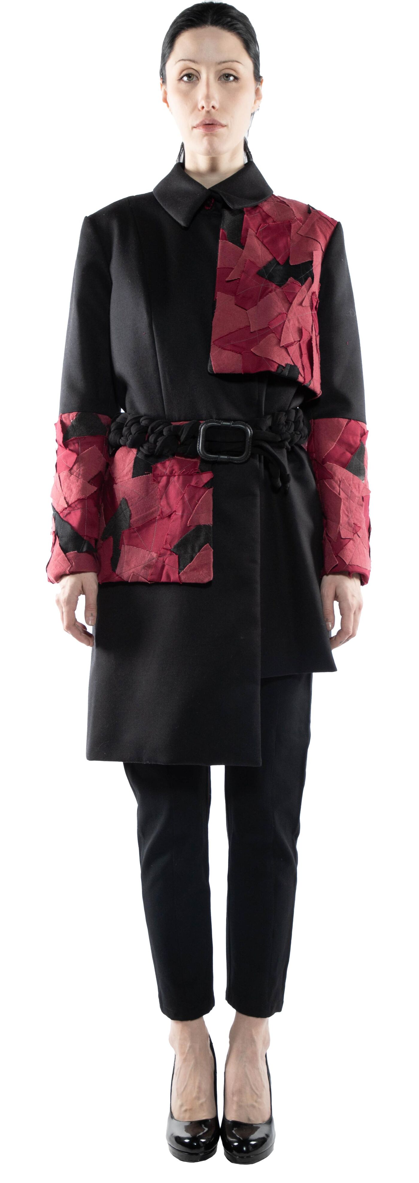 A woman is standing, wearing a black coat with red and black fabric detailing on the sleeves and a belt, paired with black pants and heels.