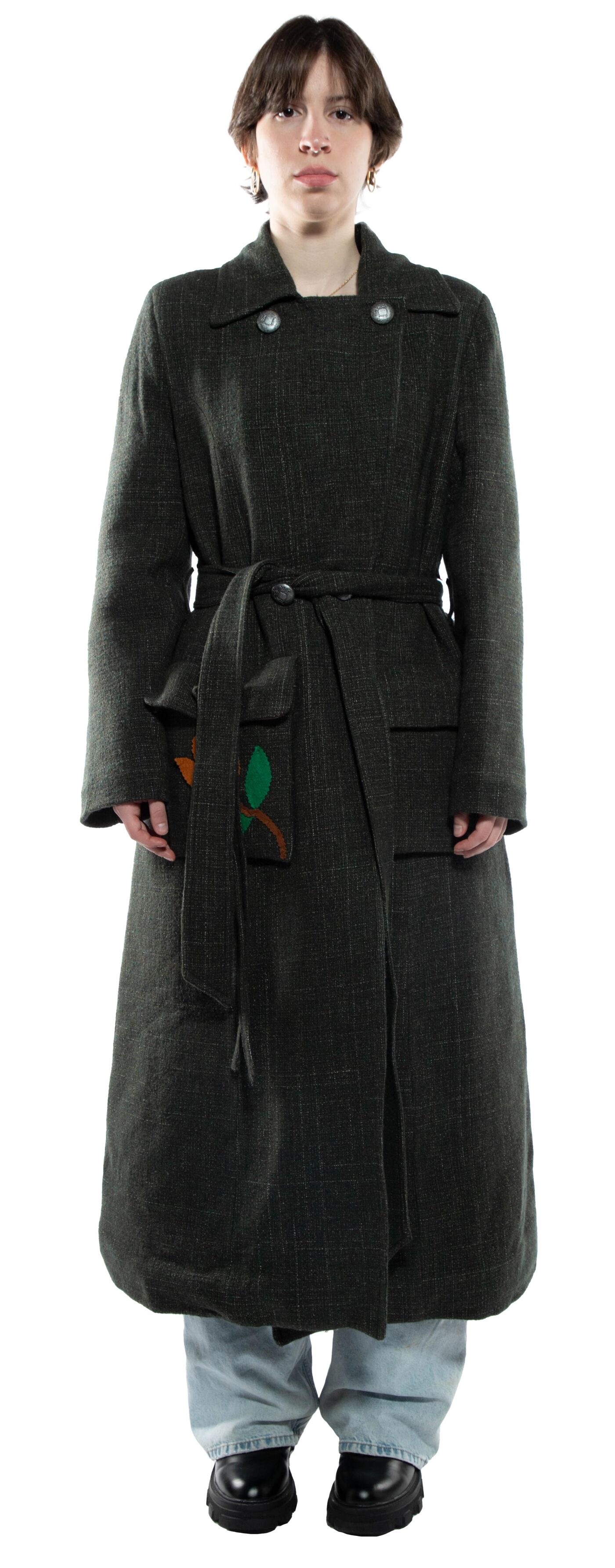 A young woman with dark hair is standing and wearing a long, dark green belted coat with pockets and light blue jeans.