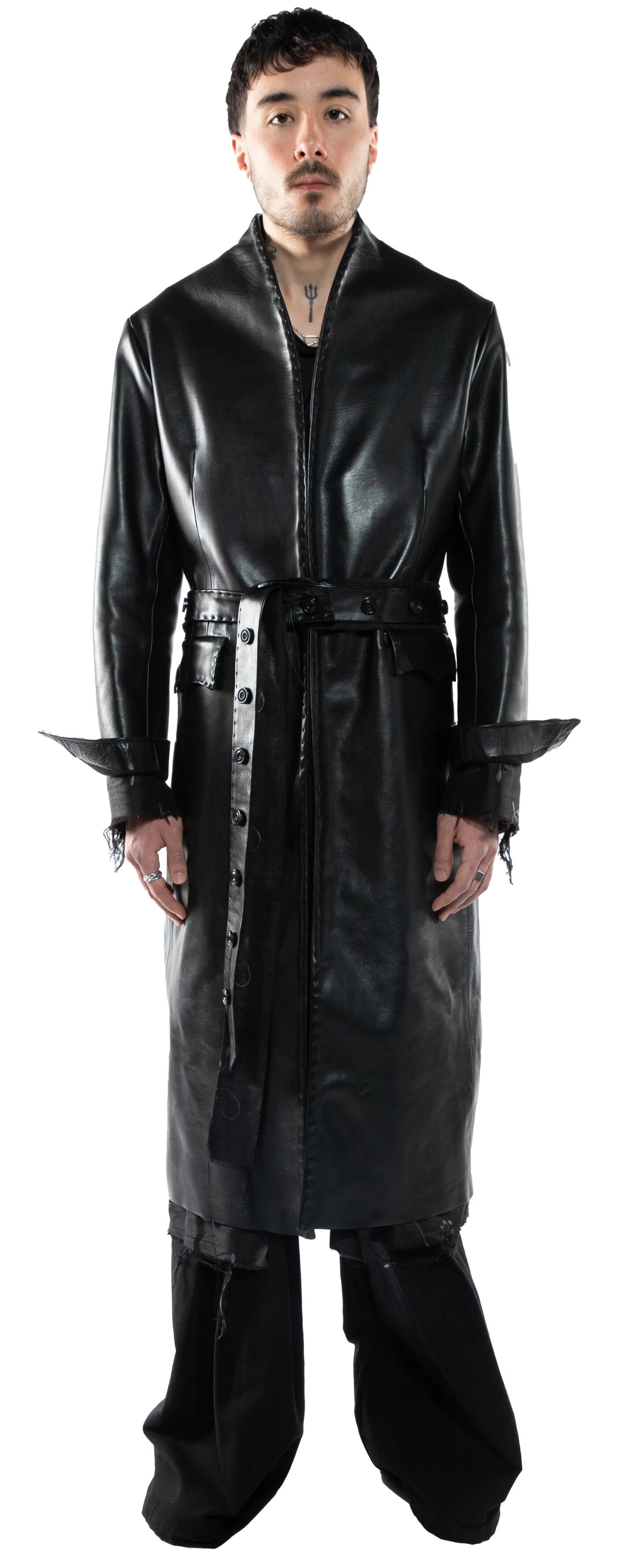 A young man with dark hair and a goatee stands against a white background, wearing a long black leather coat with a belt and matching pants, his expression is serious.