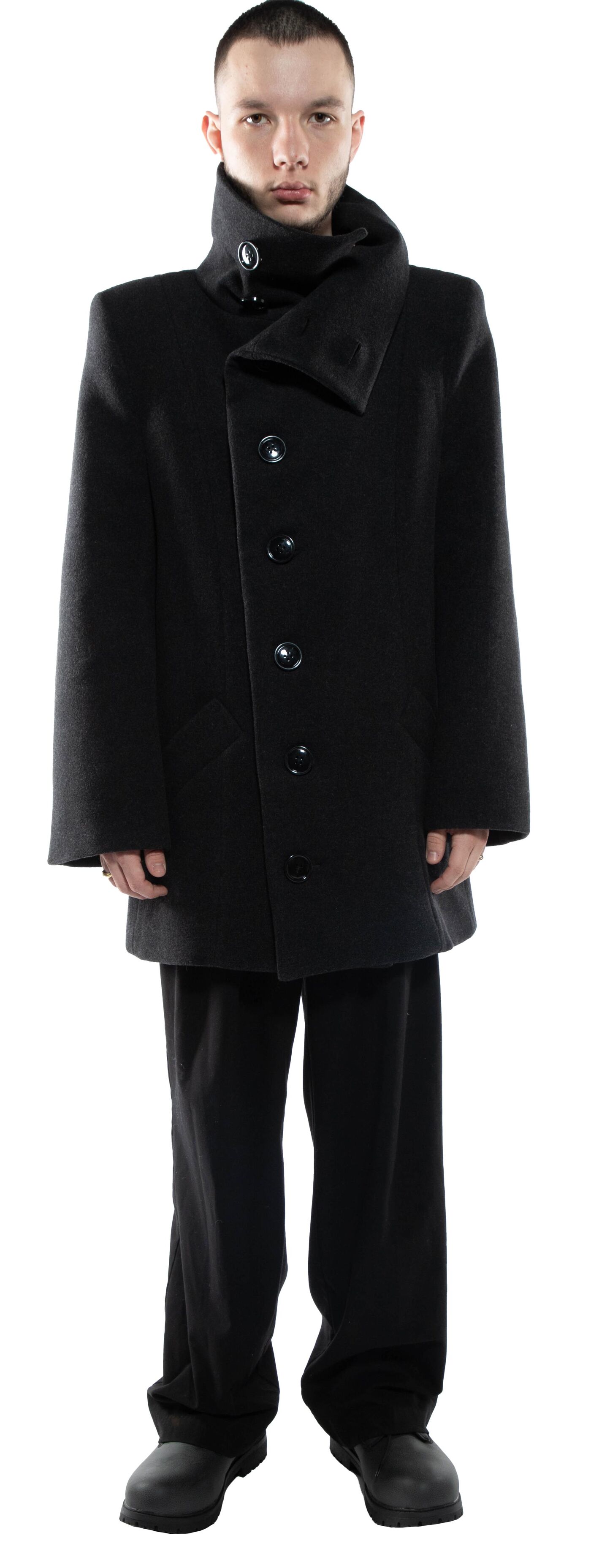 A man in a black, high-collared coat and matching trousers stands against a white background.