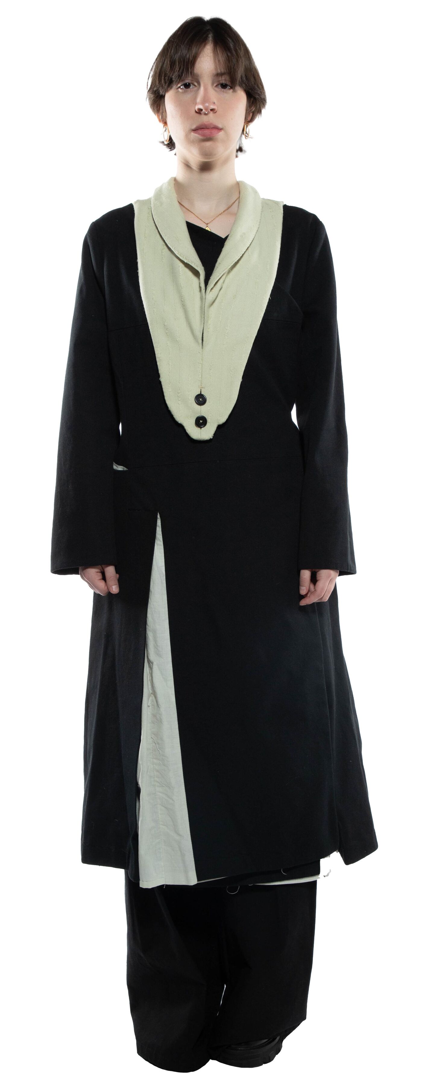 A person with short brown hair is wearing a long black and off-white coat with a high collar and black pants.