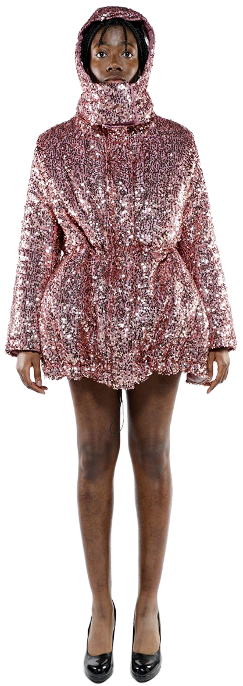 A full shot of a person of color modeling a rose-gold sequined hooded coat and black heels against a white background.