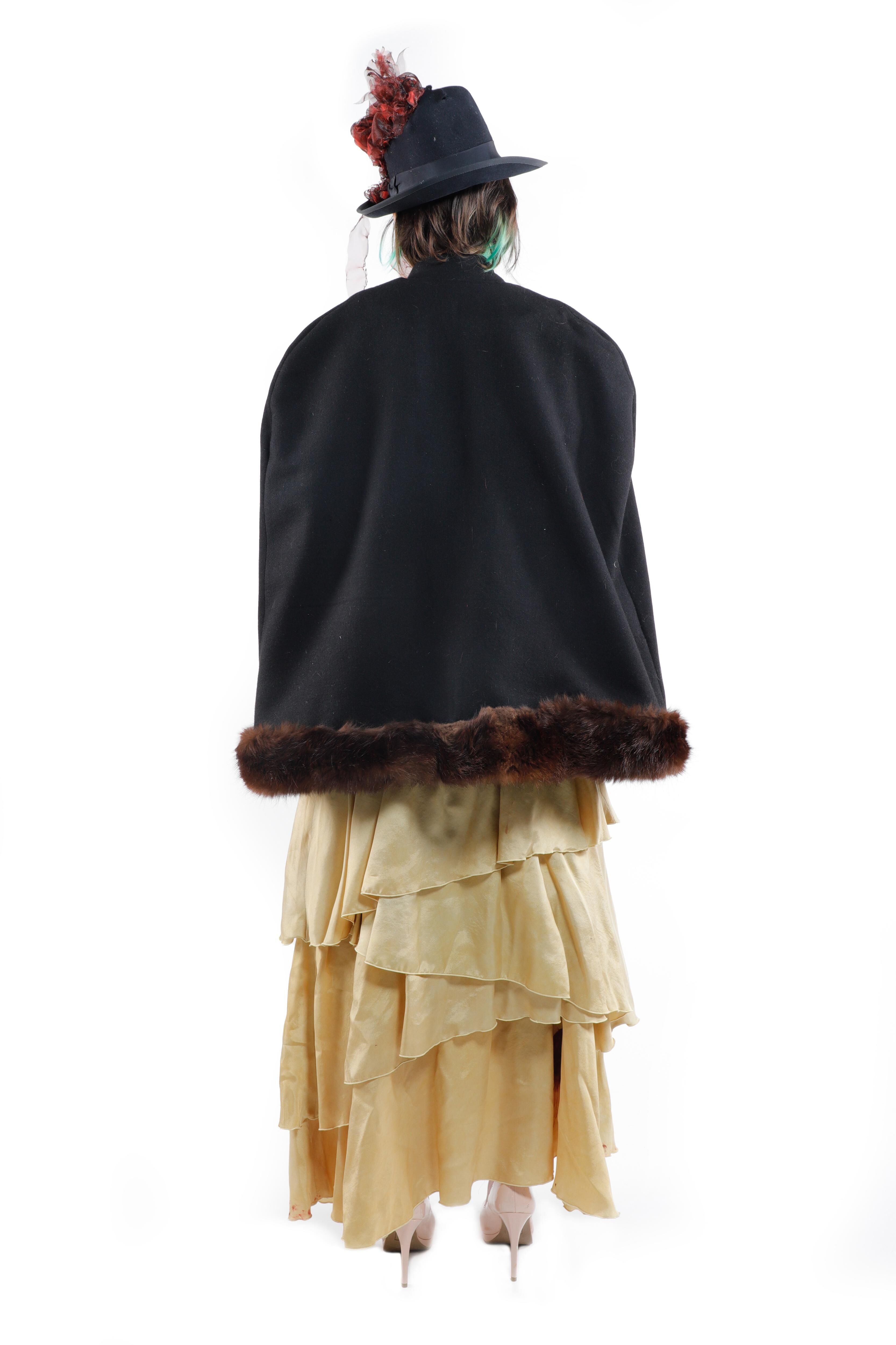 A woman wears a dark, feathered hat, a black cape with fur trim, and a long, ruffled, pale yellow skirt.
