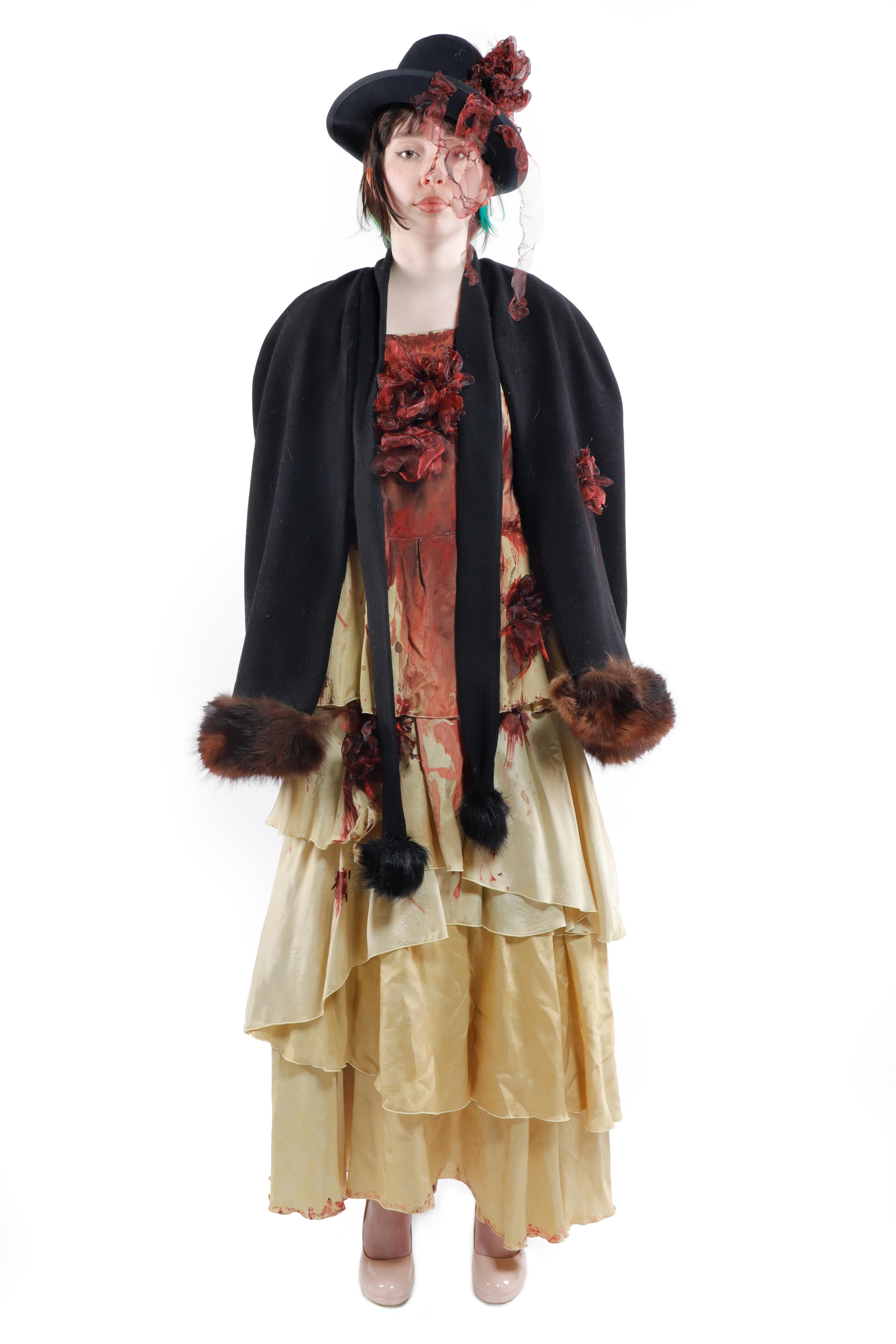 A woman wears a ruffled yellow dress, a black jacket with fur cuffs, and a black hat with a veil. The dress has red embellishments.