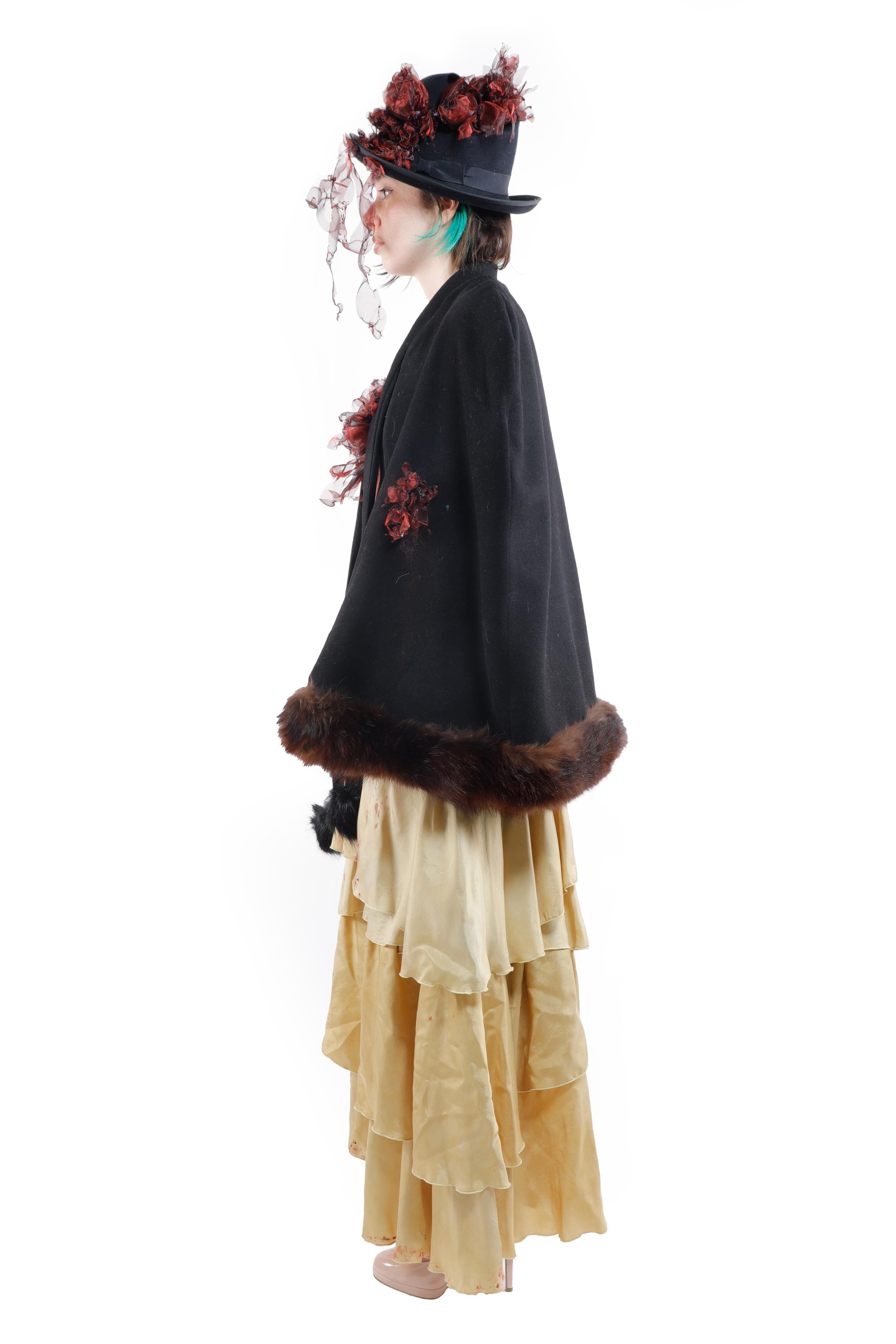 A woman wearing an ornate hat and a dark cape with fur trim, paired with a long, ruffled, golden skirt. The attire appears to be vintage.
