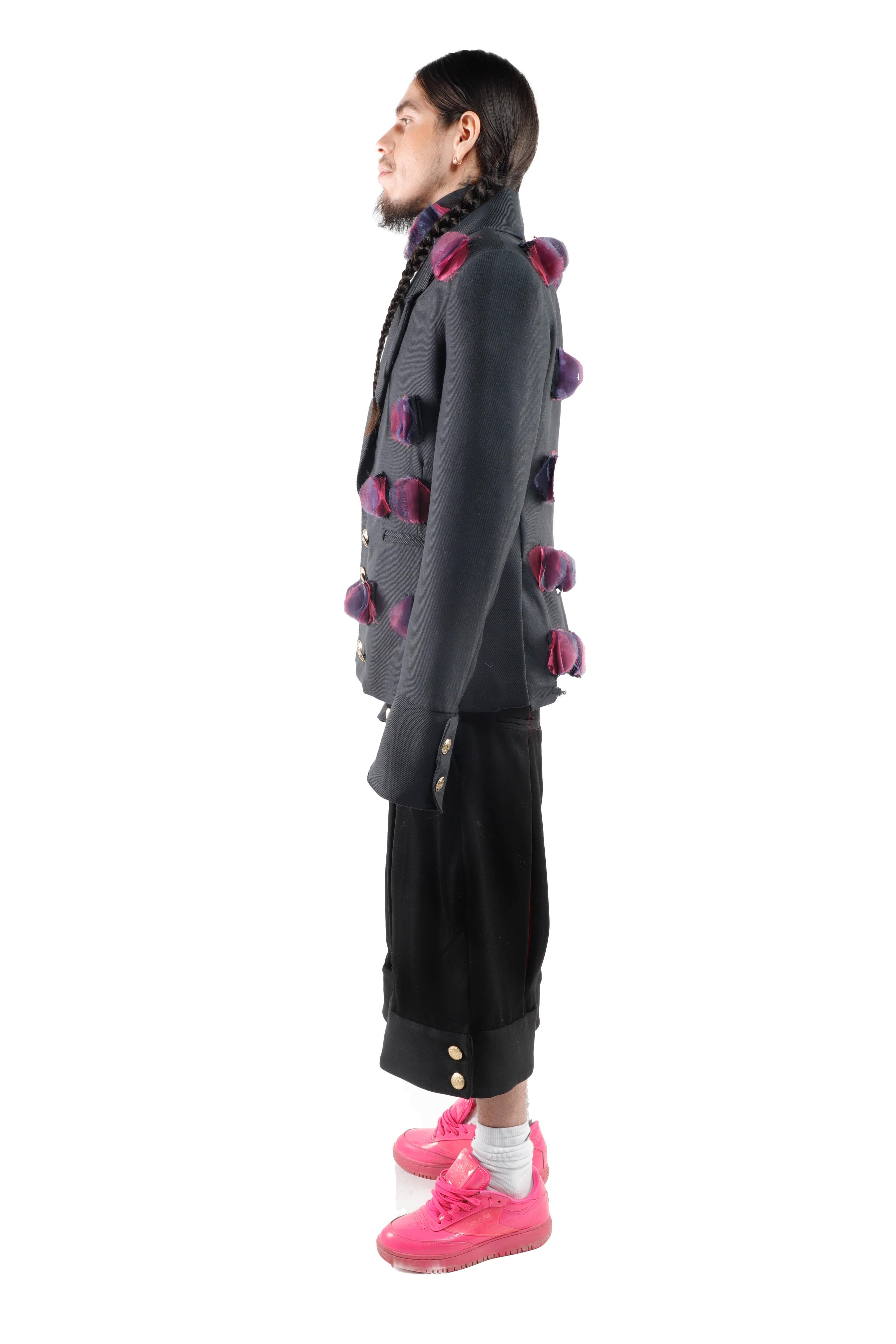 This man is wearing a dark jacket adorned with purple pom-pom details, black trousers, and vibrant pink sneakers, showcasing a bold and fashionable aesthetic.