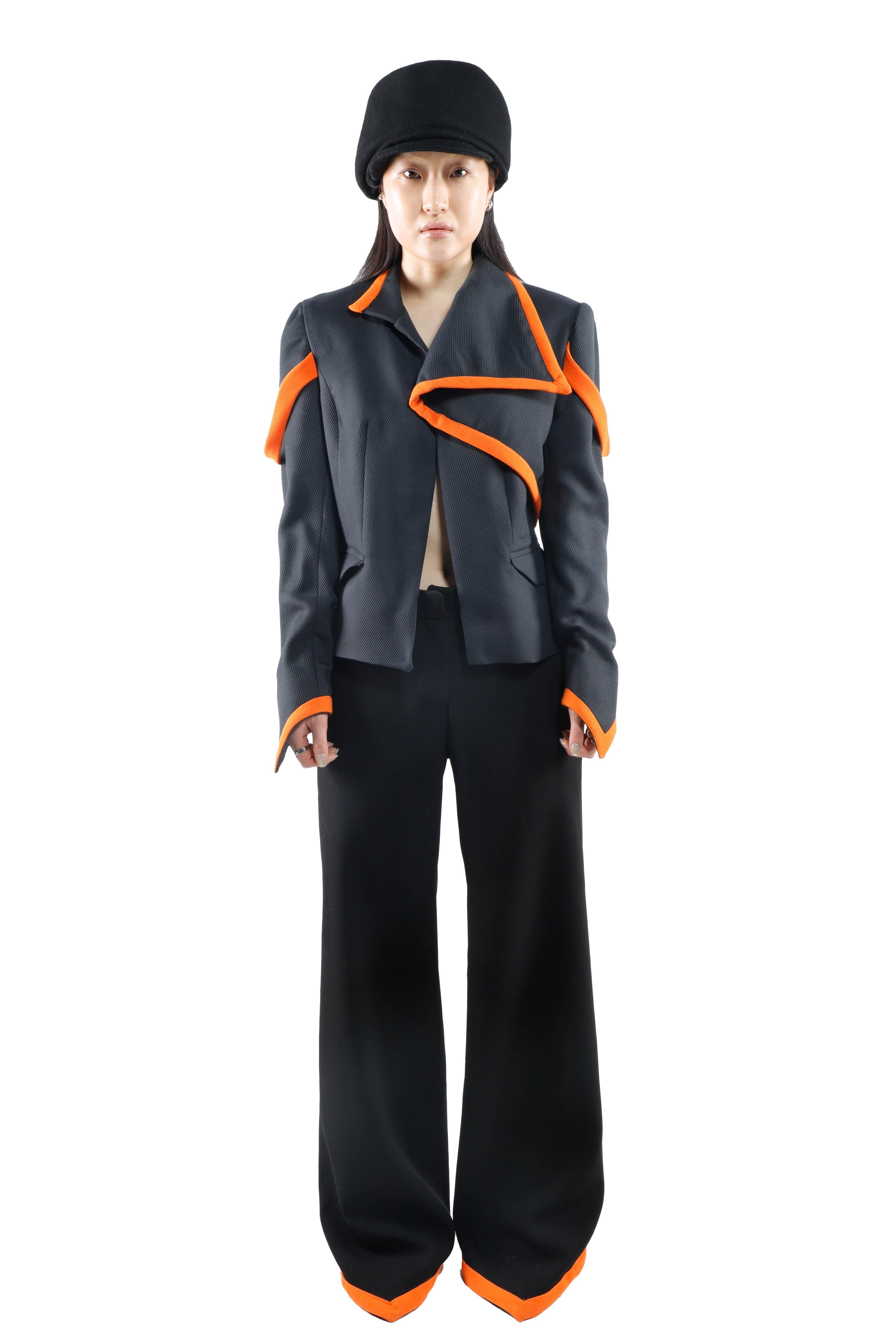 A woman wears a gray cropped jacket with orange trim and wide black pants, complemented by a black hat. The outfit features an avant-garde design.