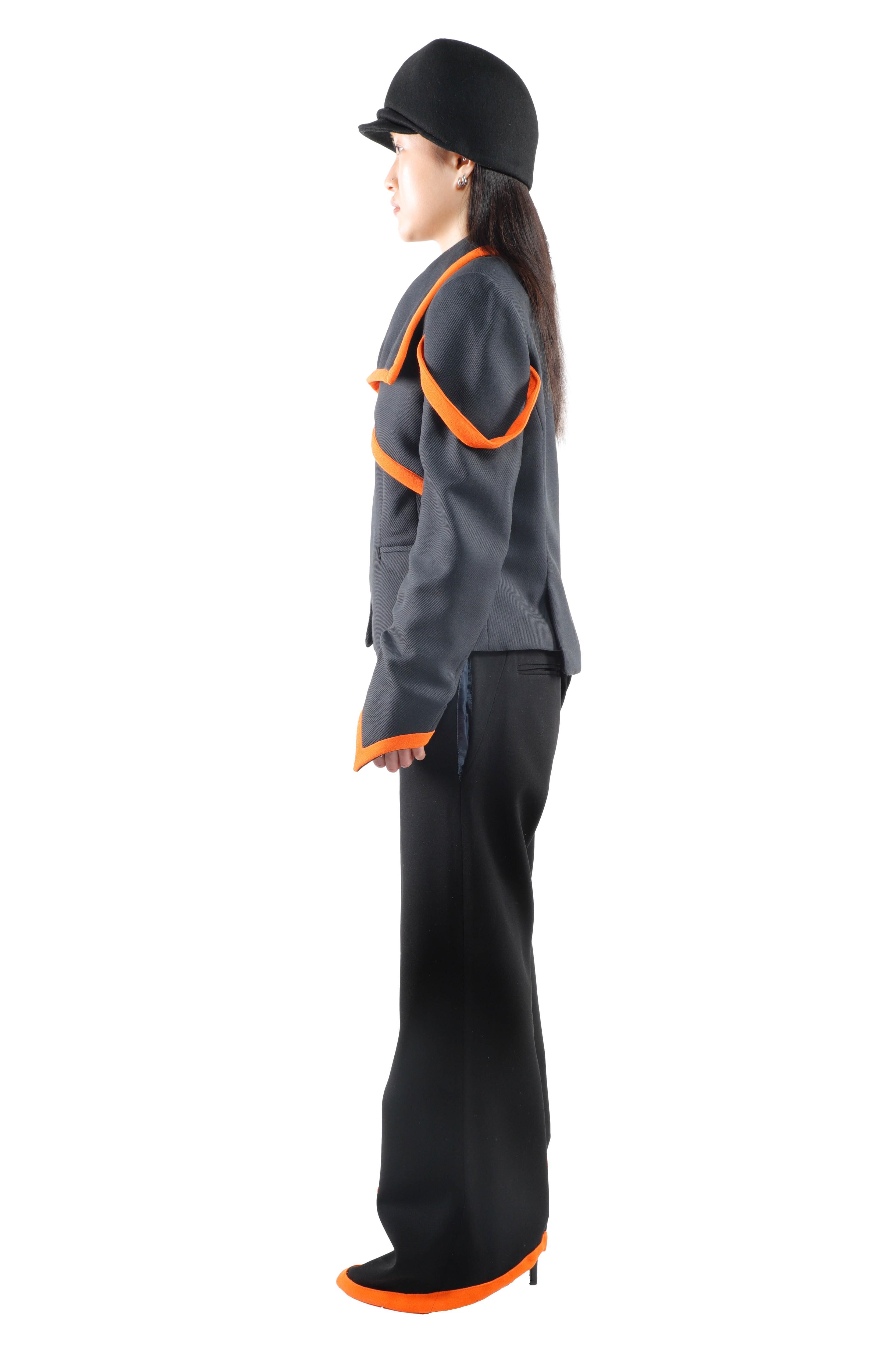 A young woman wearing a black hat and a grey outfit with orange accents, including wide-leg black pants and black shoes with orange details.