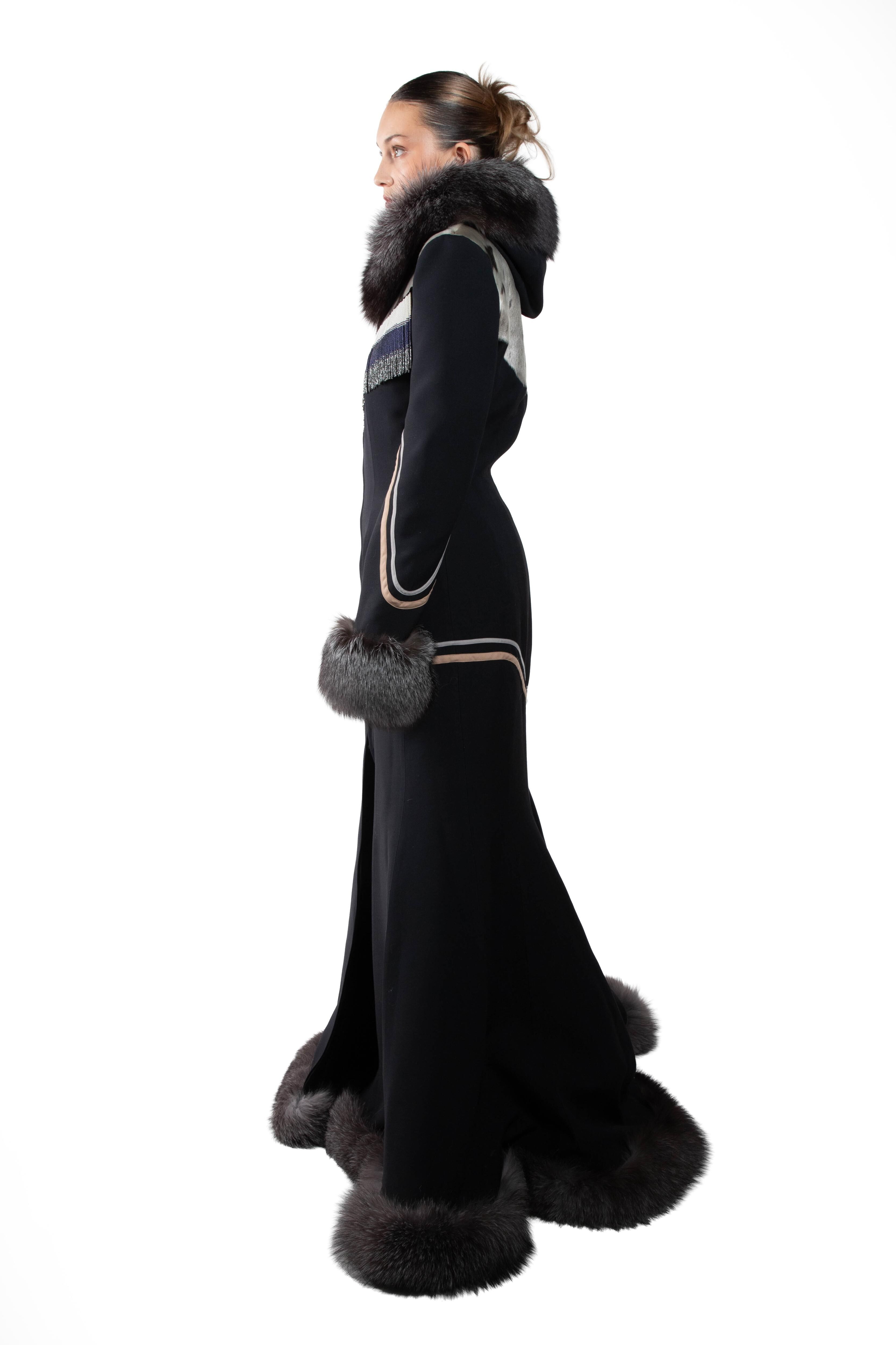 A young woman models a long black coat with grey fur accents on the collar, cuffs, and hem. The coat features an elegant design with decorative stripes on the sleeves and chest.