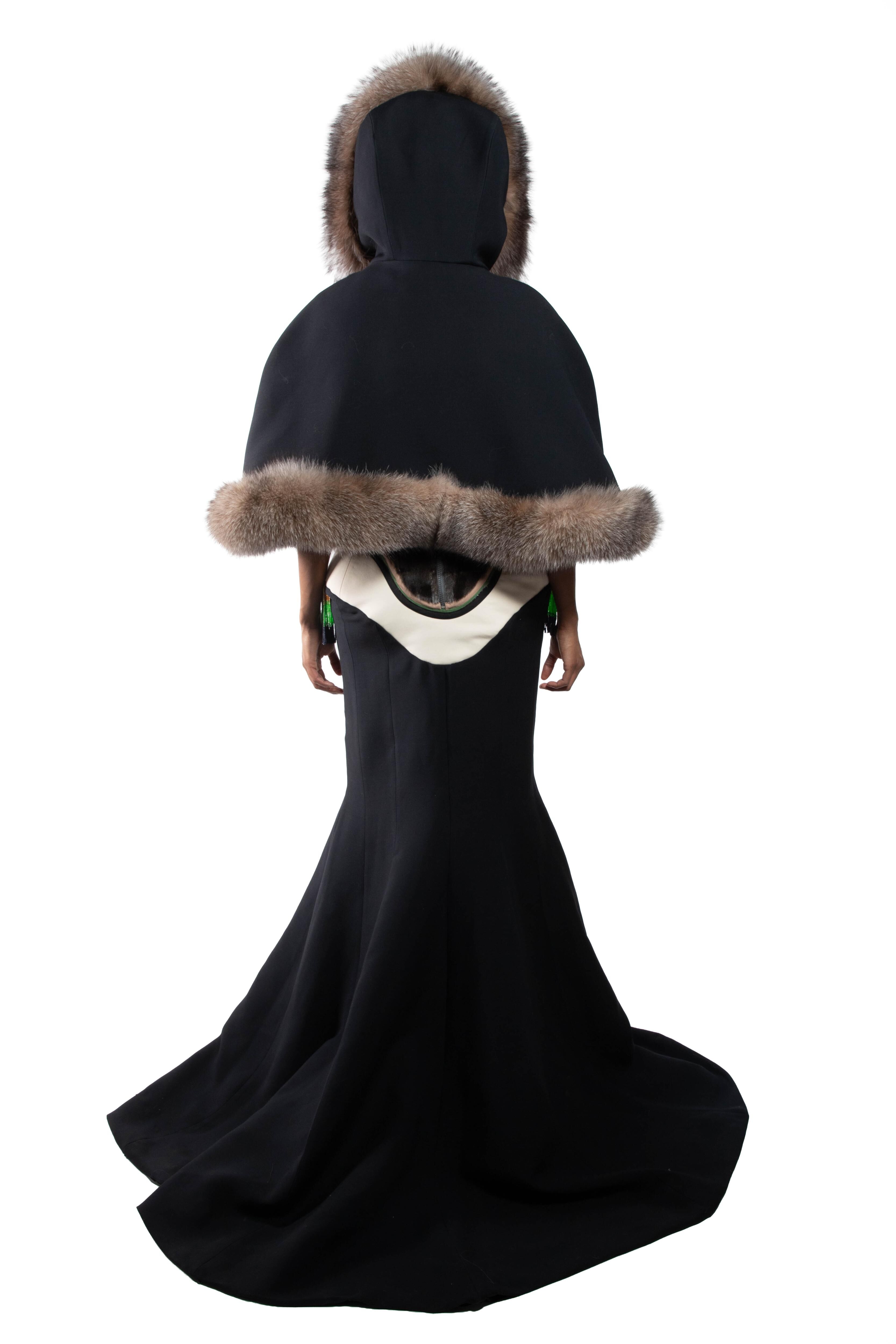 A woman wears a long black gown with a short, hooded cape trimmed with fur, creating an elegant and dramatic look.