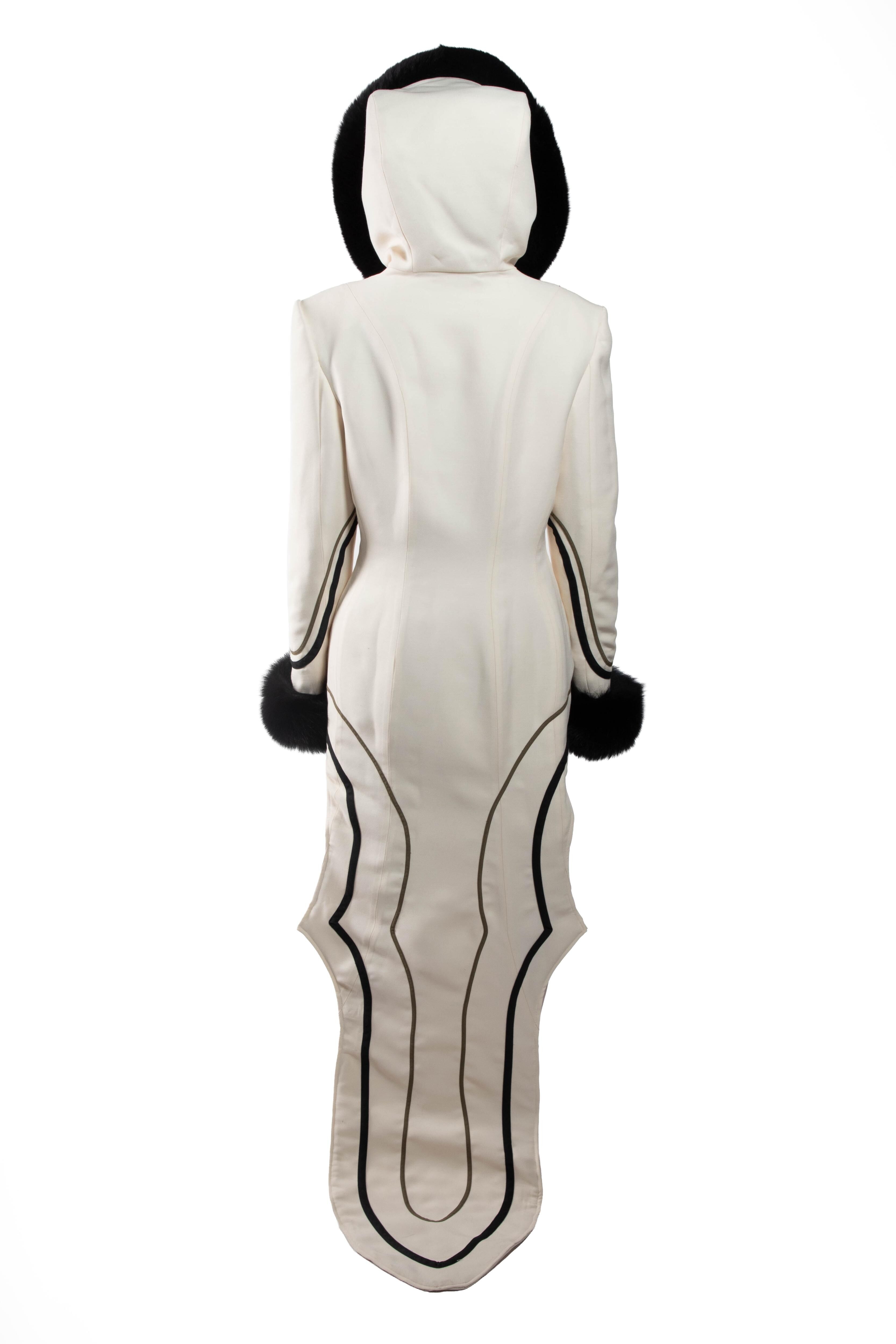 This is an image of a long white coat with black detailing, including a hood and fur cuffs, featuring a unique design at the bottom.