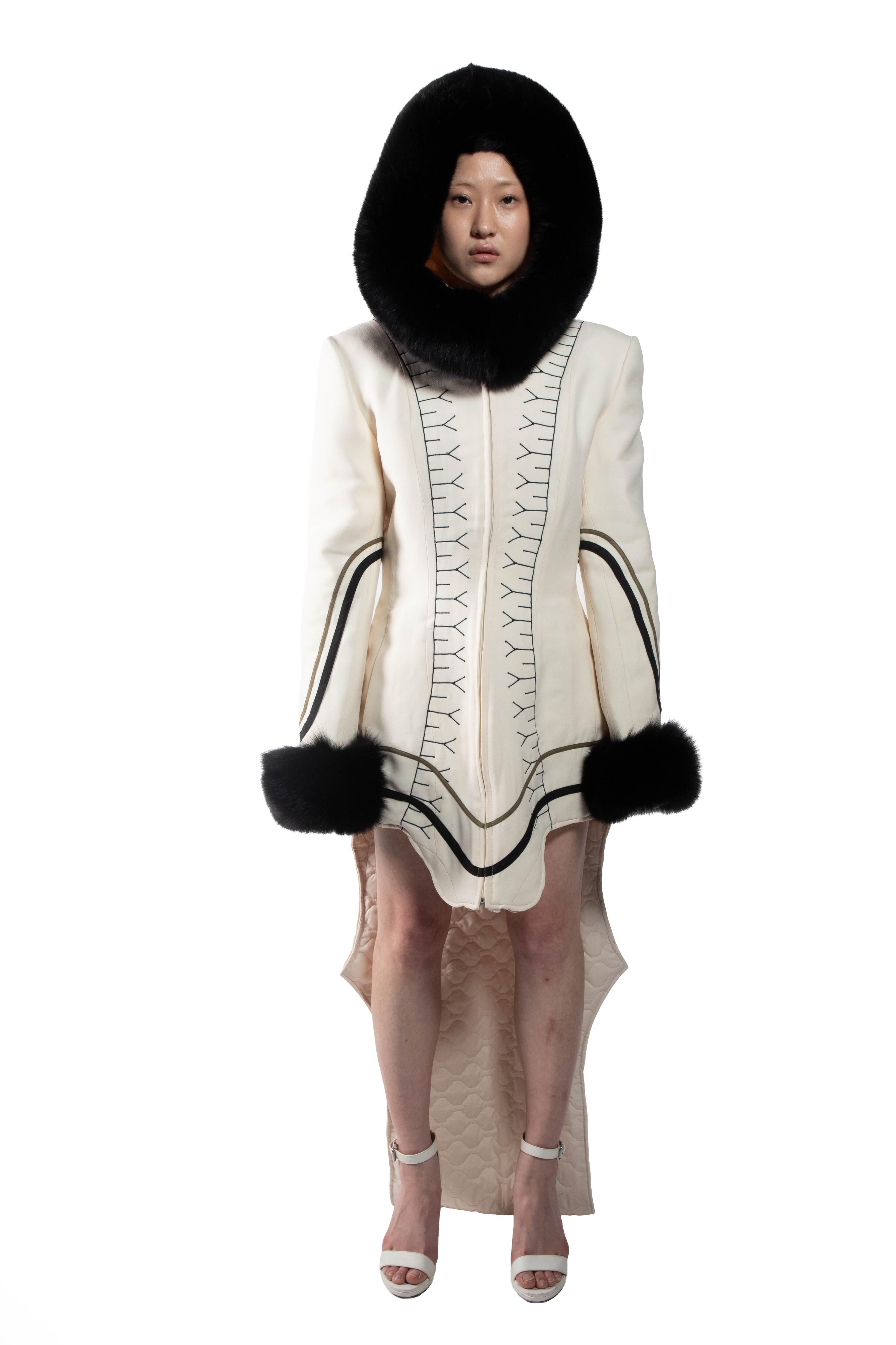 A woman is featured wearing a long, white coat with black detailing and a black fur hood, complemented by white high-heeled shoes.