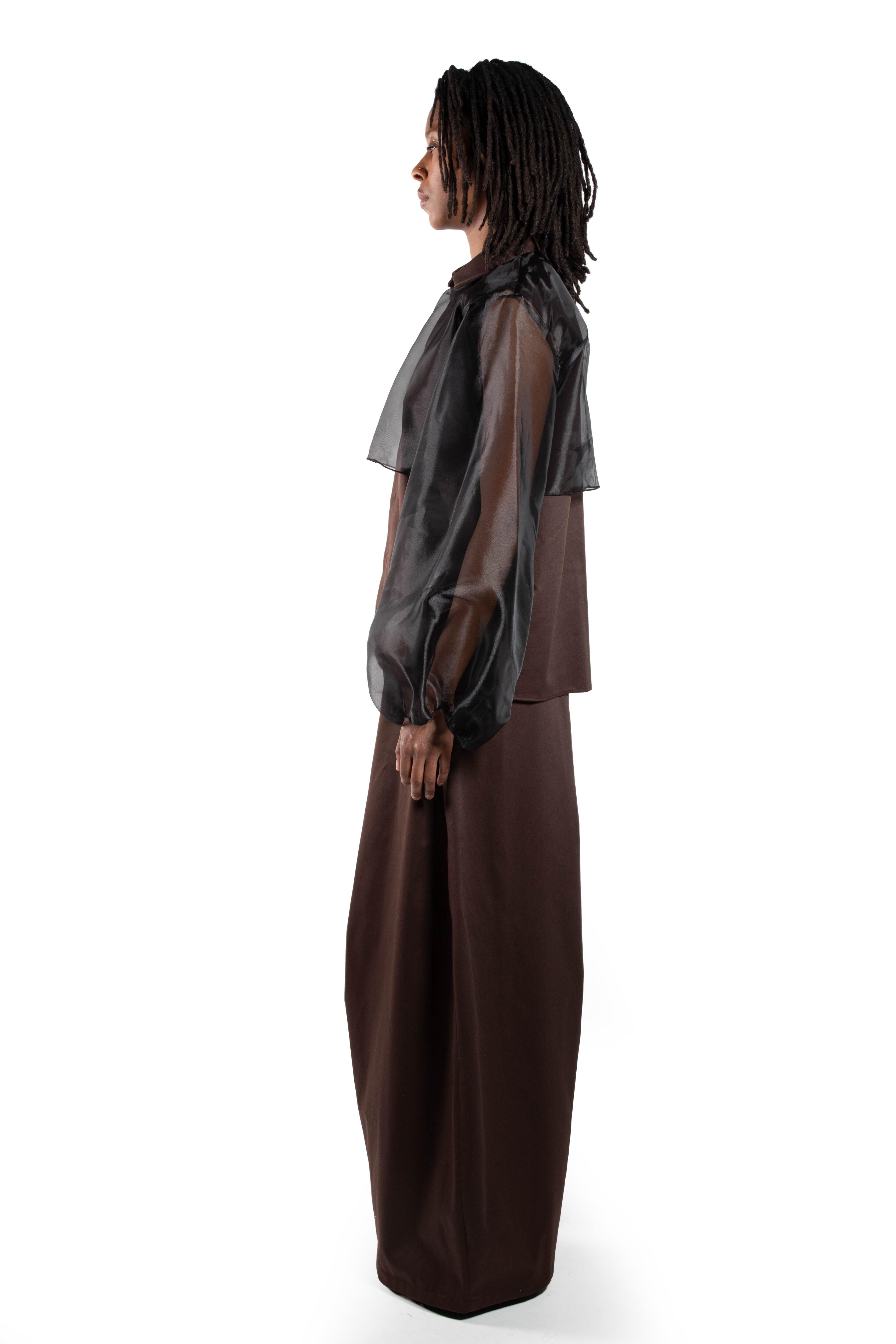 A woman with dreadlocks is wearing a sheer, long-sleeved blouse and a long brown skirt. The image is taken from the side against a white background.