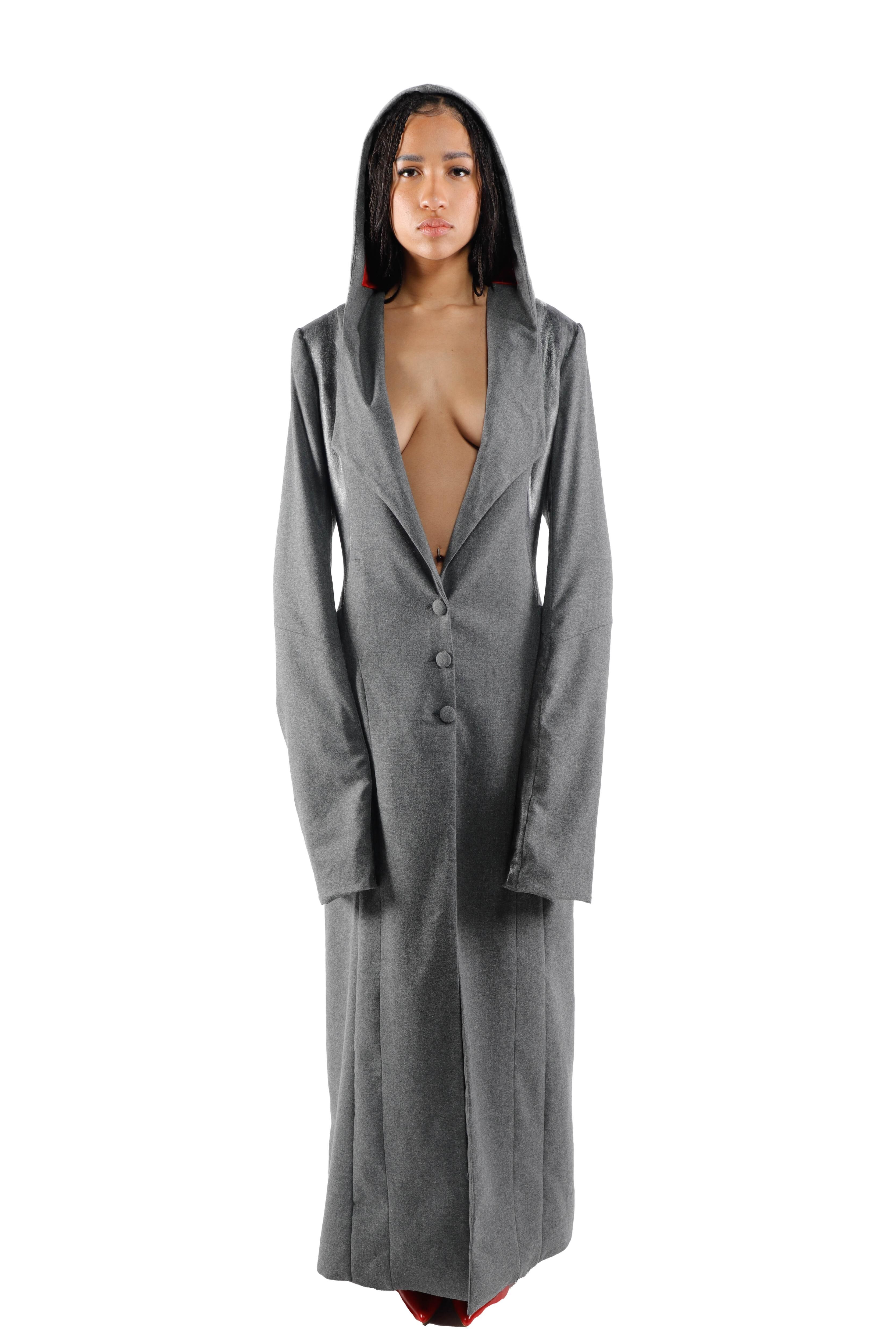 A young woman with long, dark hair is wearing a long, gray coat with a deep V-neckline. She has a serious expression as she looks directly at the camera.