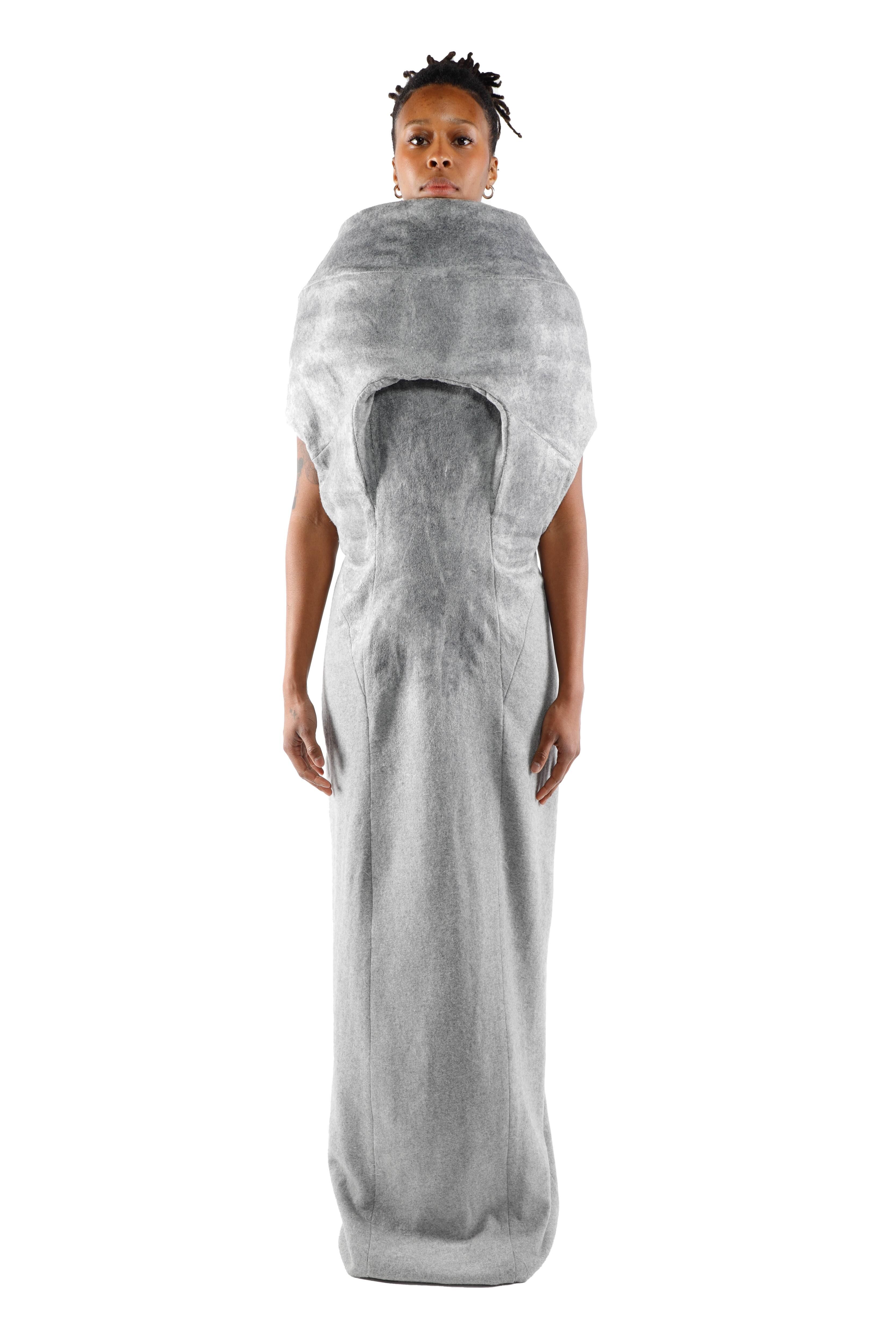 A woman wears a long, gray dress with a sculptural, voluminous design around the neck and shoulders, creating a unique silhouette.