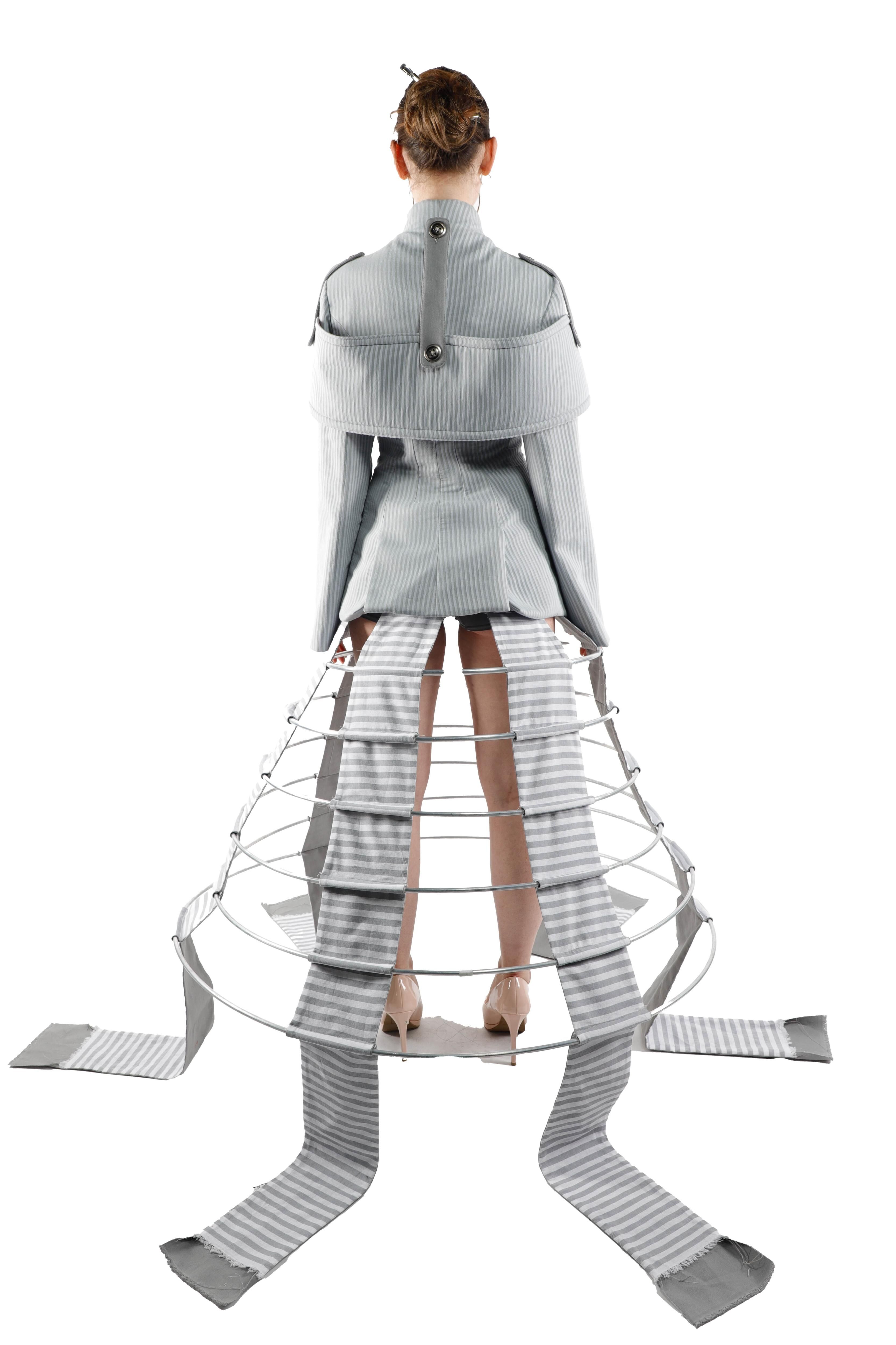 This fashion design features a woman in a light gray outfit with a metallic structure extending from the waist, adorned with striped fabric strips.