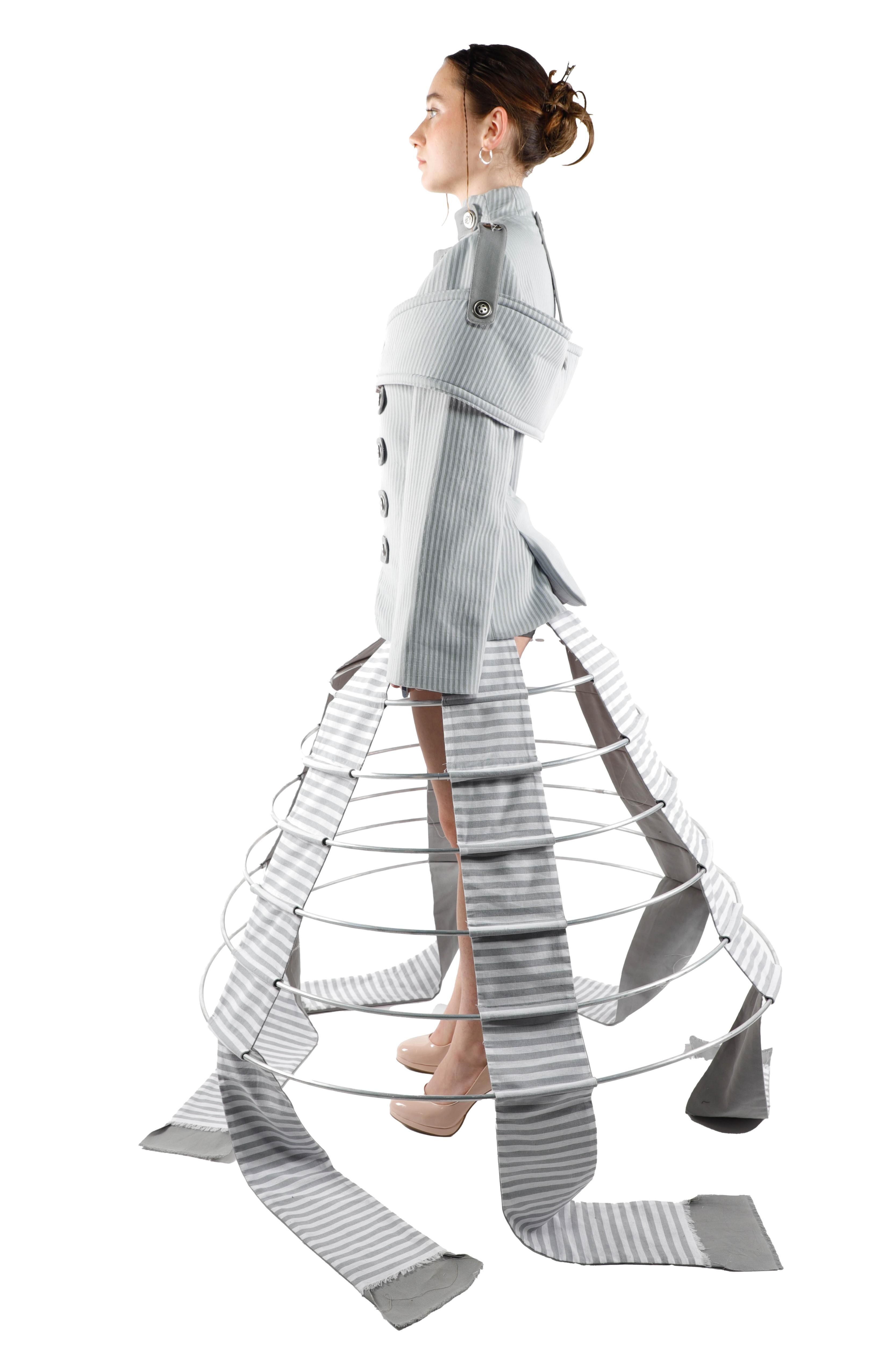 A young woman models a conceptual outfit featuring a pale grey jacket and a skirt with a cage-like structure adorned with striped fabric strips.