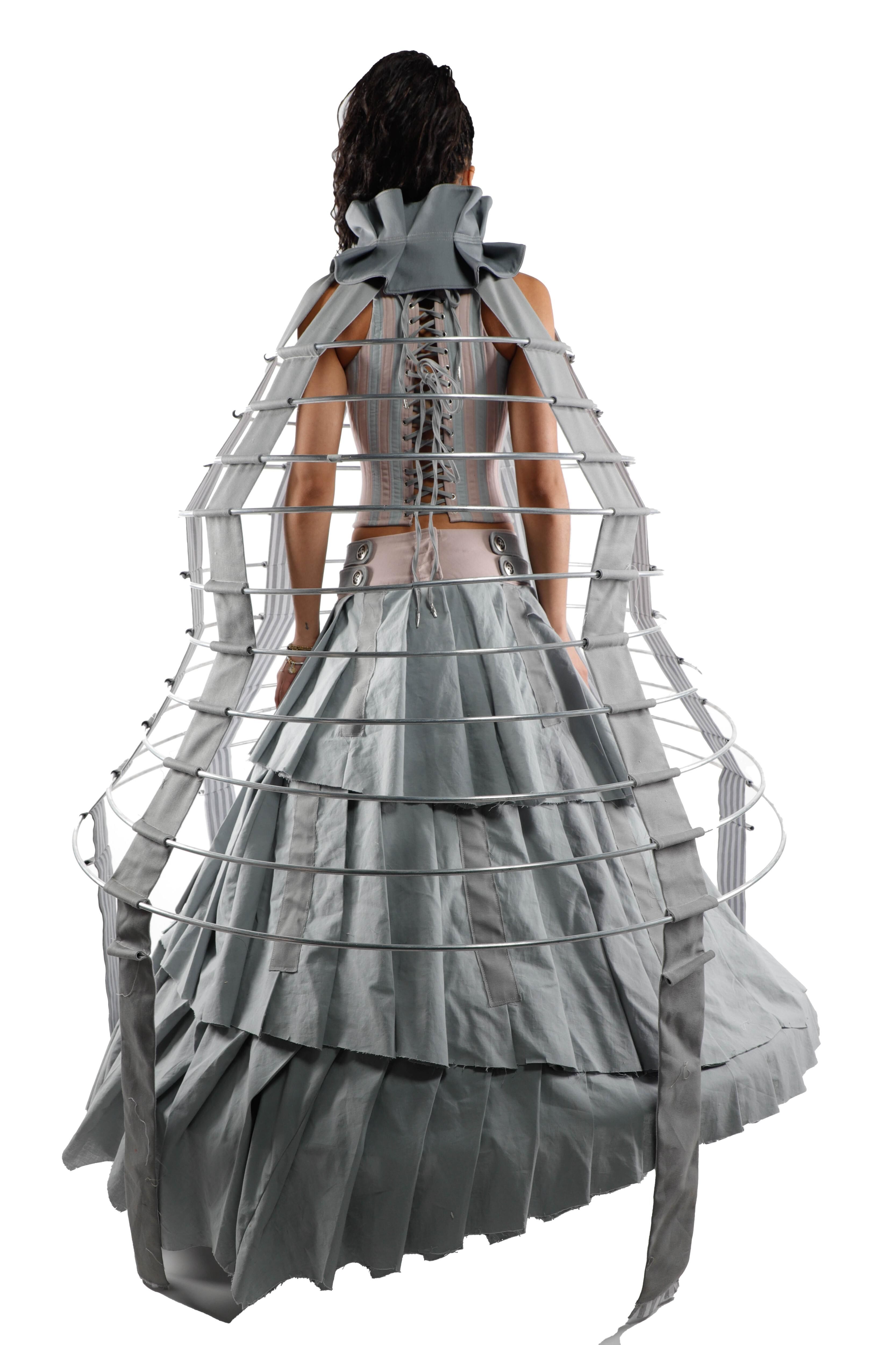 This image shows a woman viewed from the back, wearing a gray dress with a cage-like structure that creates a voluminous and dramatic silhouette.