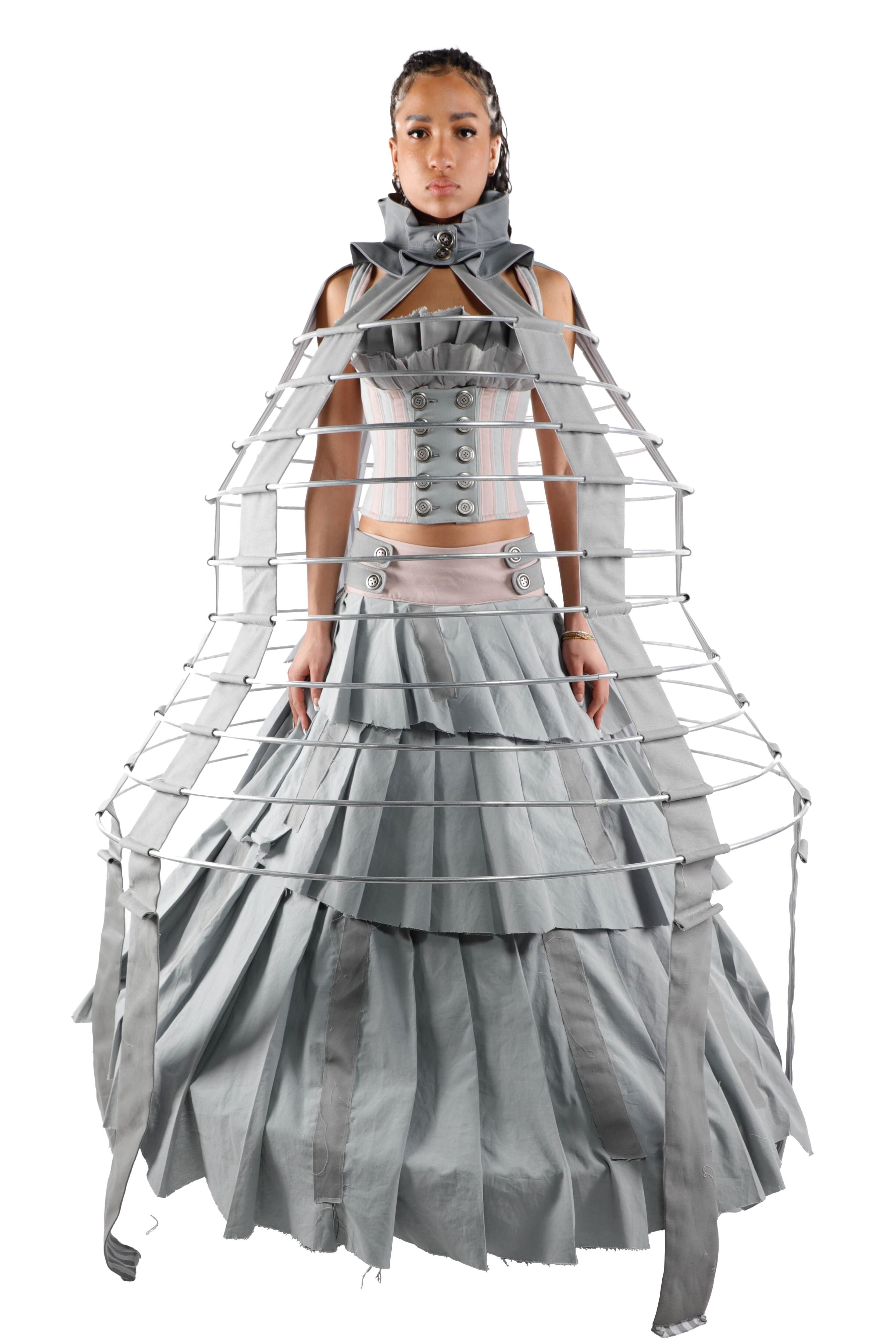 A woman wears a gray dress with a metal structure extending from the waist, creating a dramatic and avant-garde silhouette.