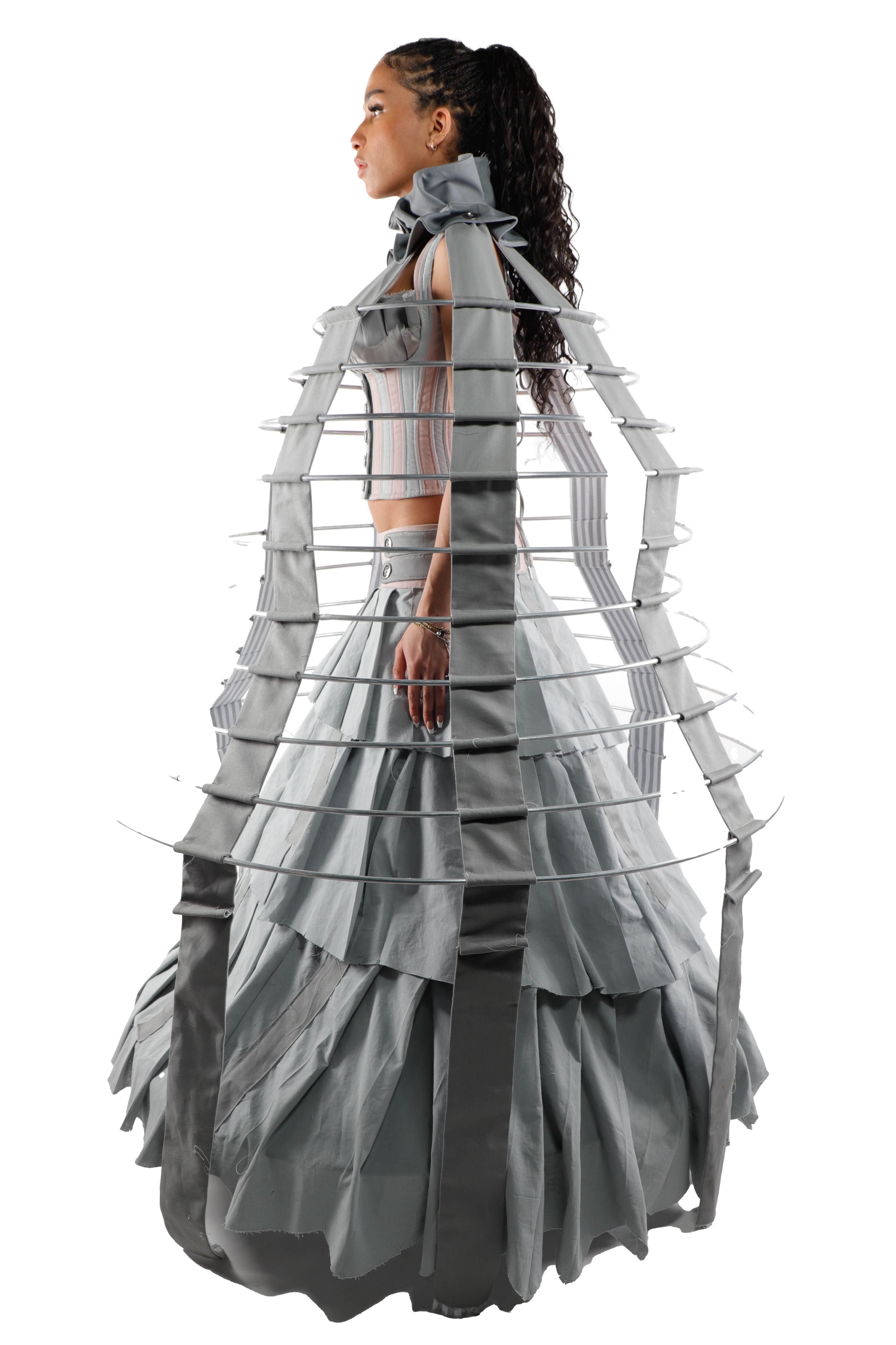 A young woman with dark hair in a ponytail wears a gray dress with a cage-like structure under the skirt. The dress features ruffles and a high neckline.