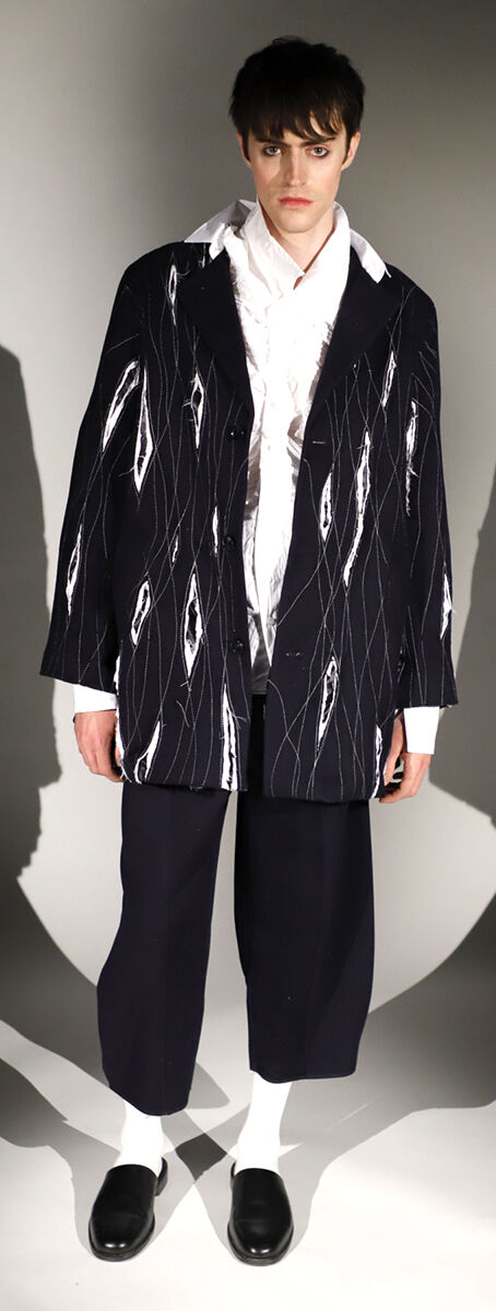 A young man models a black jacket with a white abstract print, a white shirt, and wide-leg black pants, paired with black shoes and white socks.