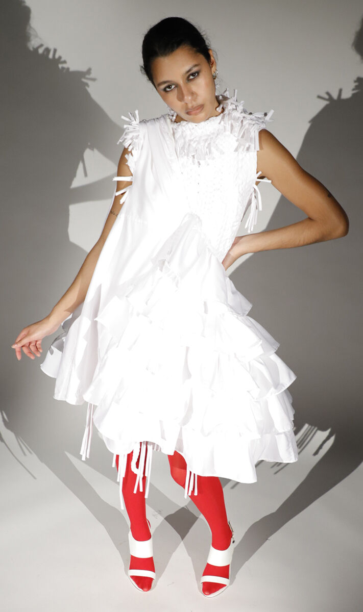 A young woman wearing red stockings and red and white shoes is modeling a white dress with ruffled layers and fringe details.