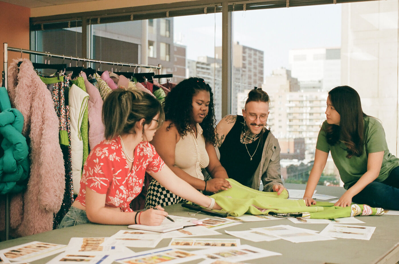 A diverse group of four individuals focused on designing garments, surrounded by colorful fabric and sketches in a creative studio setting.