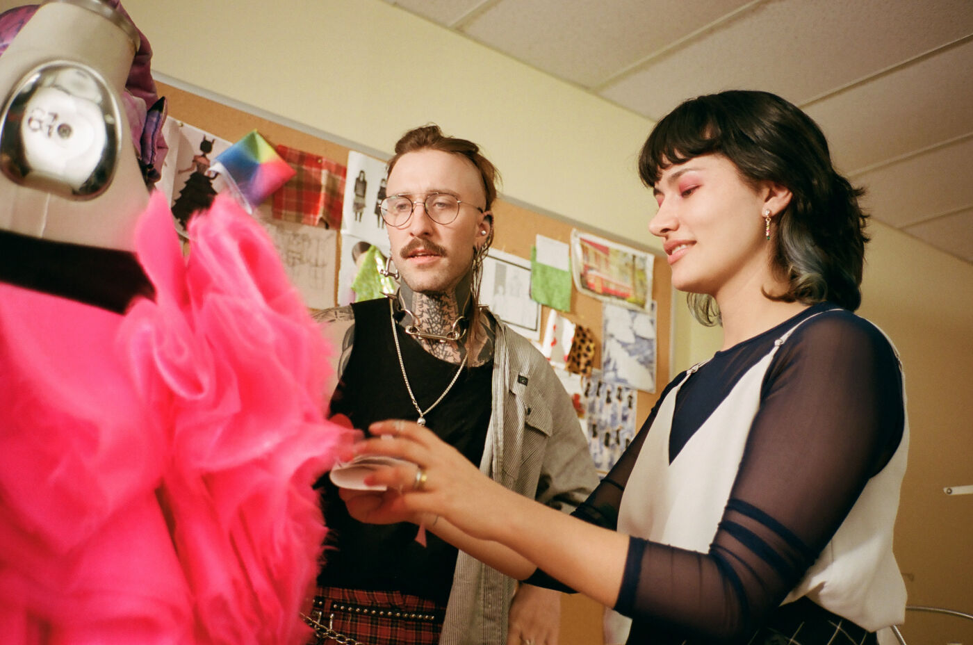 Two smiling fashion designers work on a vibrant pink garment on a mannequin in a studio filled with design sketches.