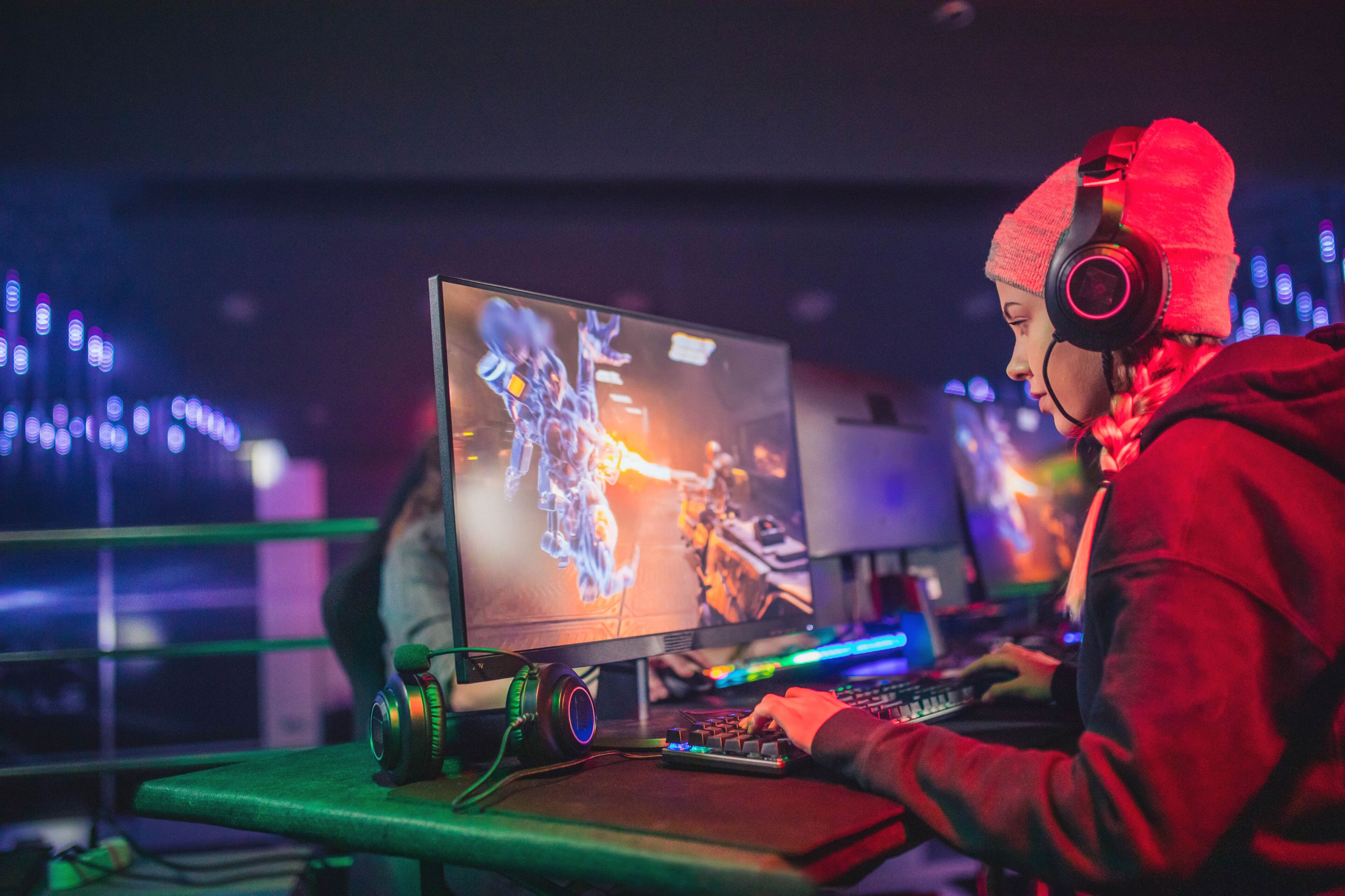 A focused gamer wearing a red beanie and headphones plays a competitive video game at an illuminated eSports arena.