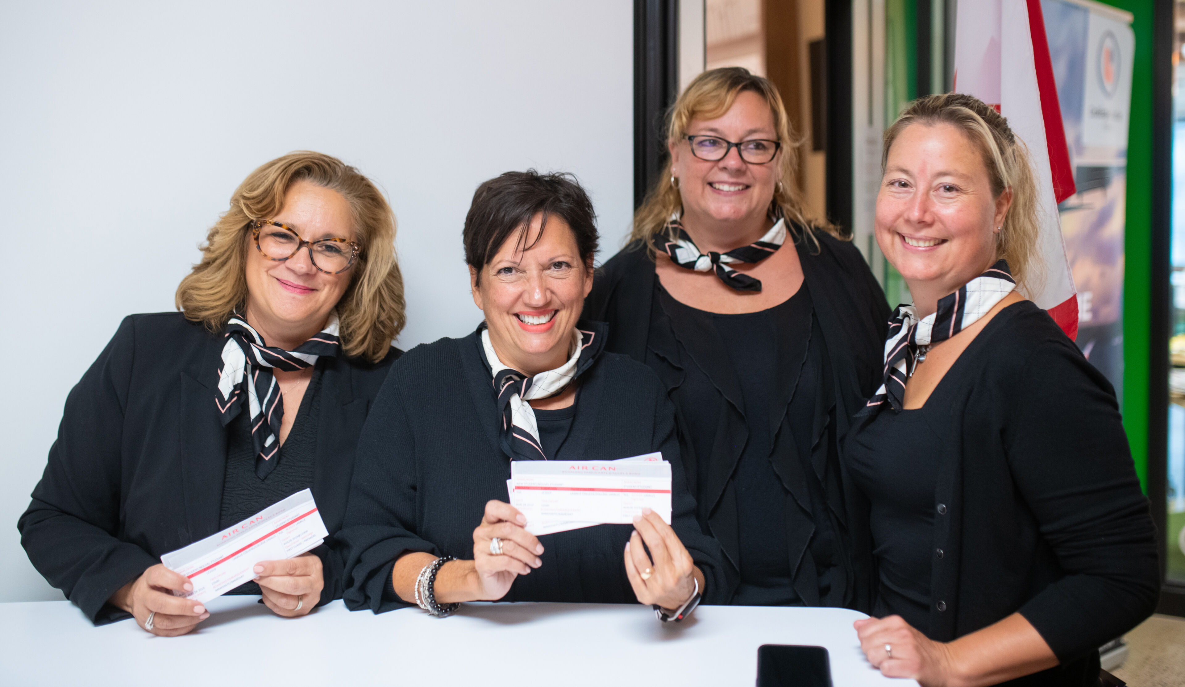 Four cheerful female hosts at a corporate event, wearing black uniforms with matching scarves, displaying event passes.