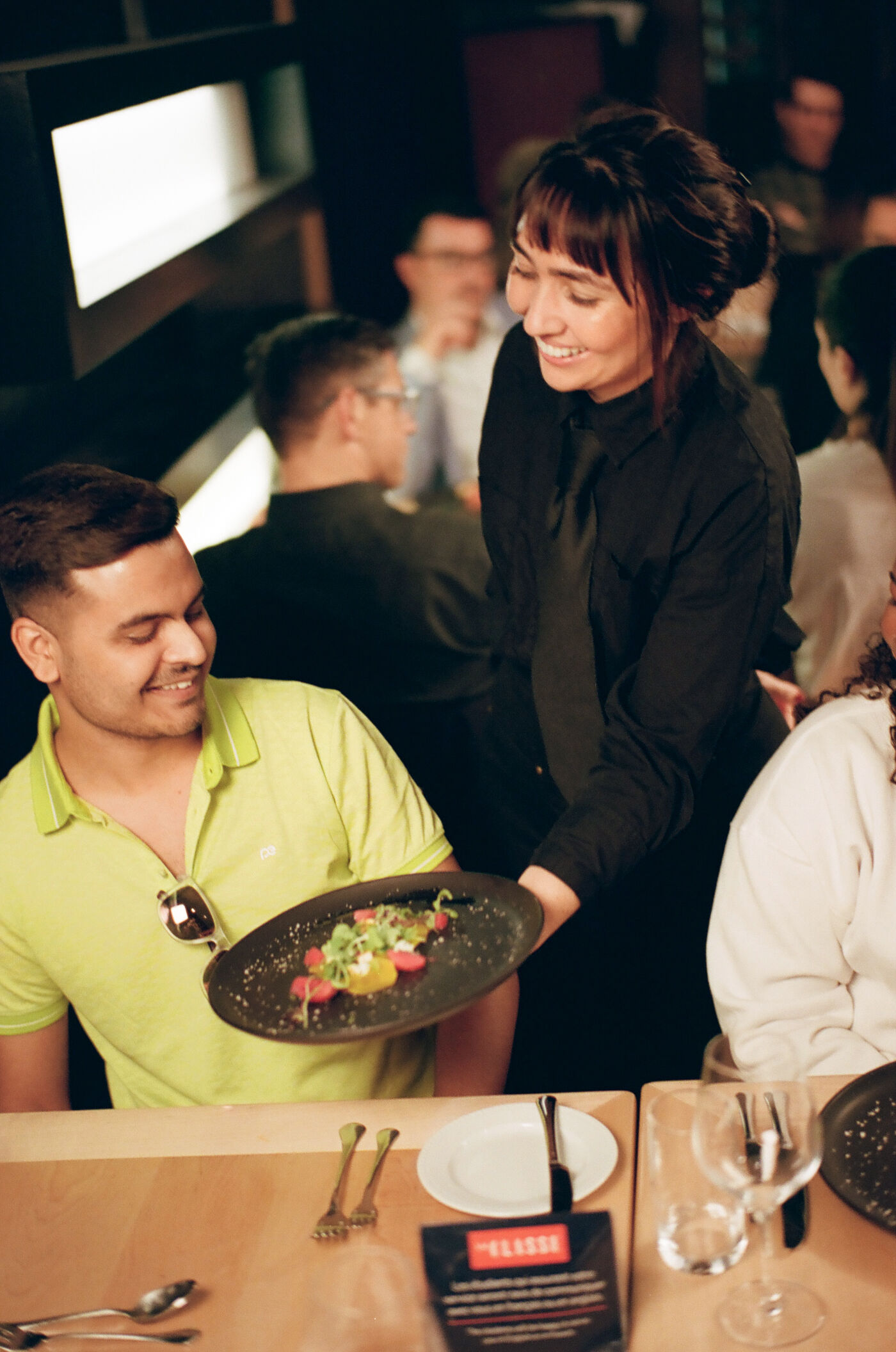 A smiling server in a black uniform presents a gourmet dish to a seated patron wearing a lime green shirt in a bustling restaurant.