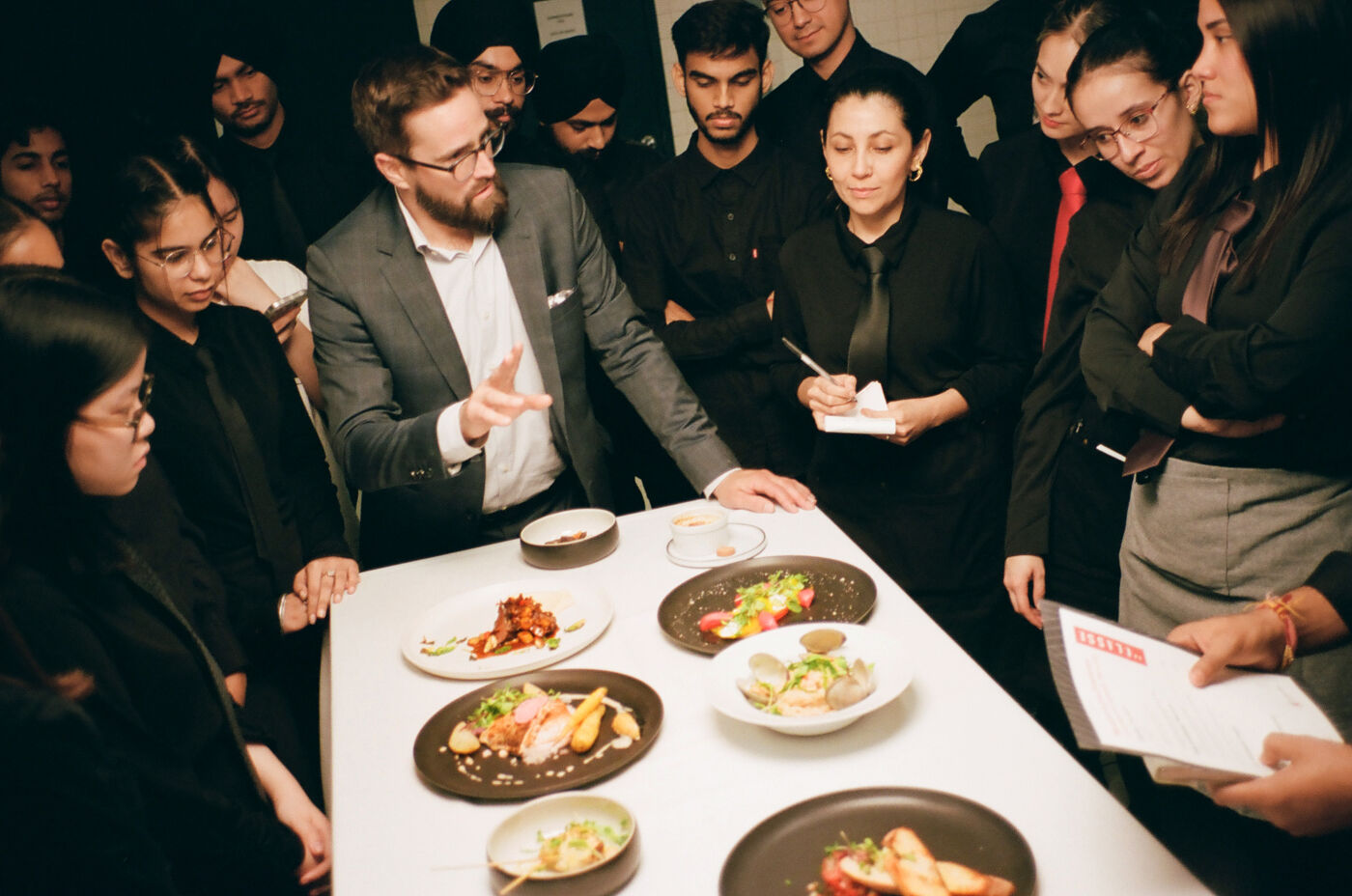 A chef explains dish preparations to attentive hospitality professionals gathered around a table with plated dishes.