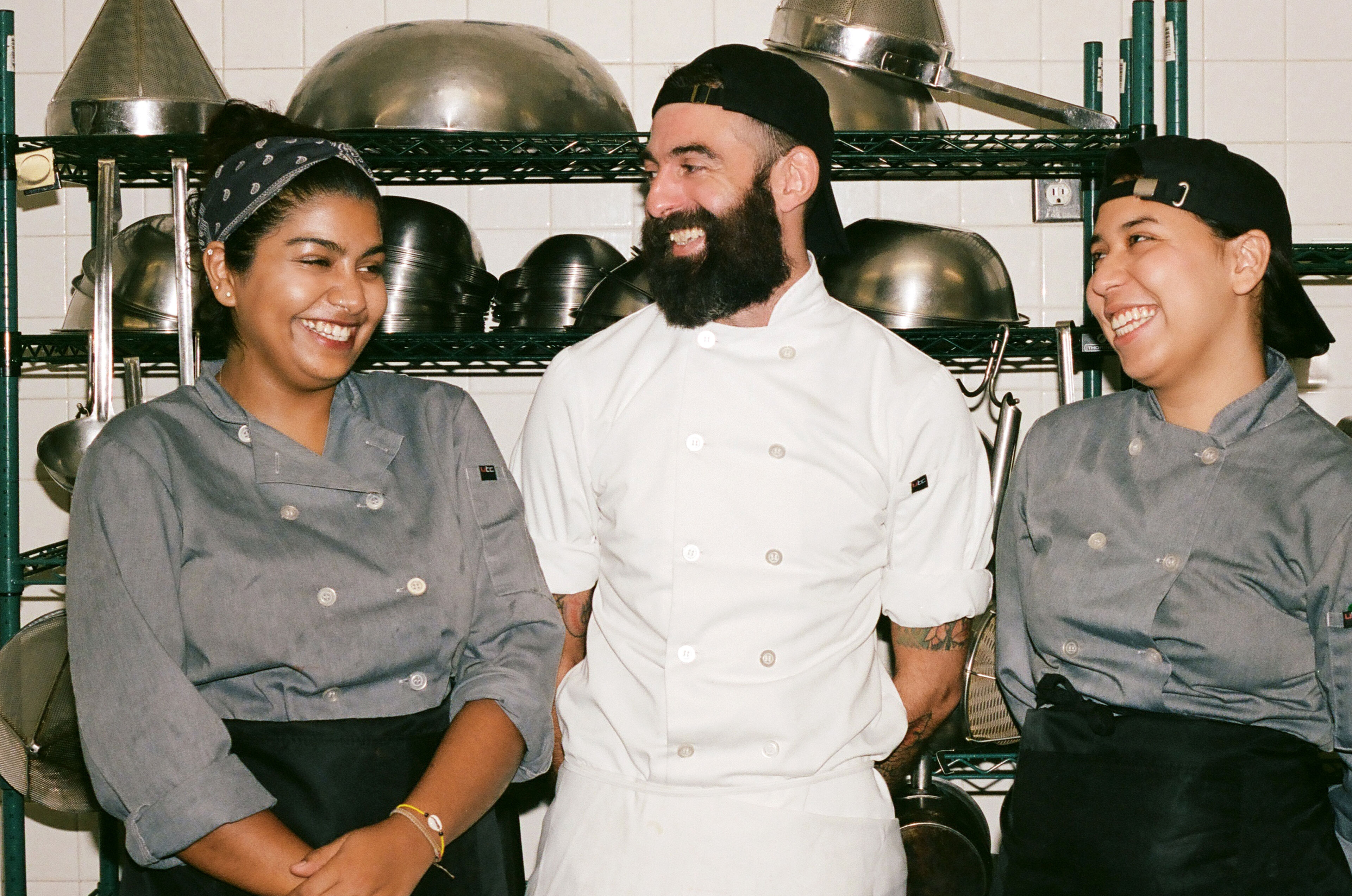 Tres cocineros compartiendo un momento alegre en una cocina comercial concurrida, con ollas de acero inoxidable y estanterías al fondo.