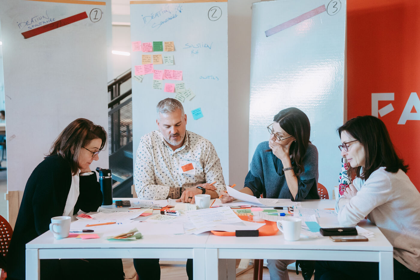 The image shows four people sitting around a table, seemingly engaged in a collaborative work session. They are surrounded by papers, markers, and sticky notes, suggesting a brainstorming or planning activity in a bright, modern office space.