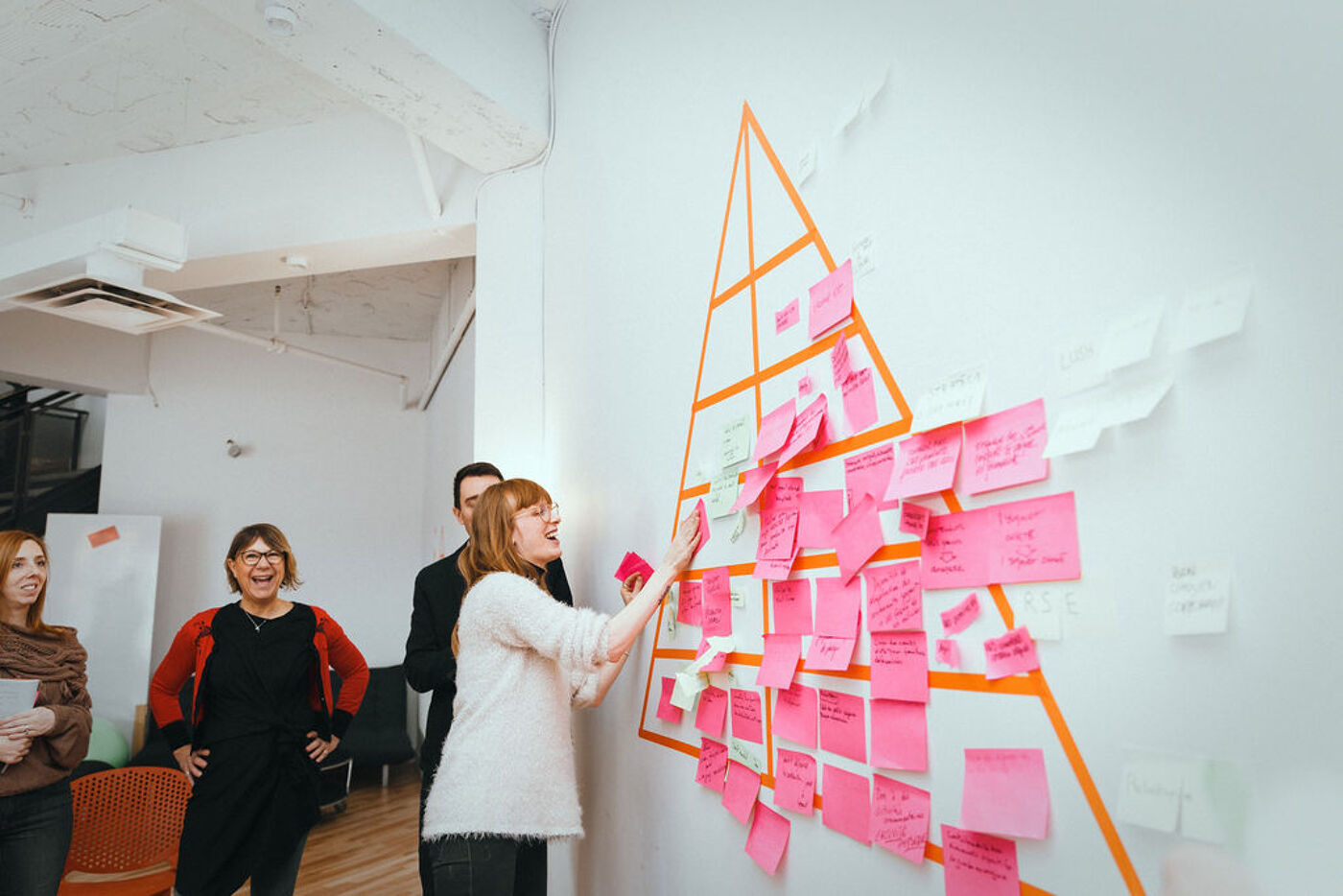 A group of people are gathered around a wall covered in pink sticky notes arranged in a pyramid shape. They appear to be brainstorming or collaborating on a project in a bright, modern office space.